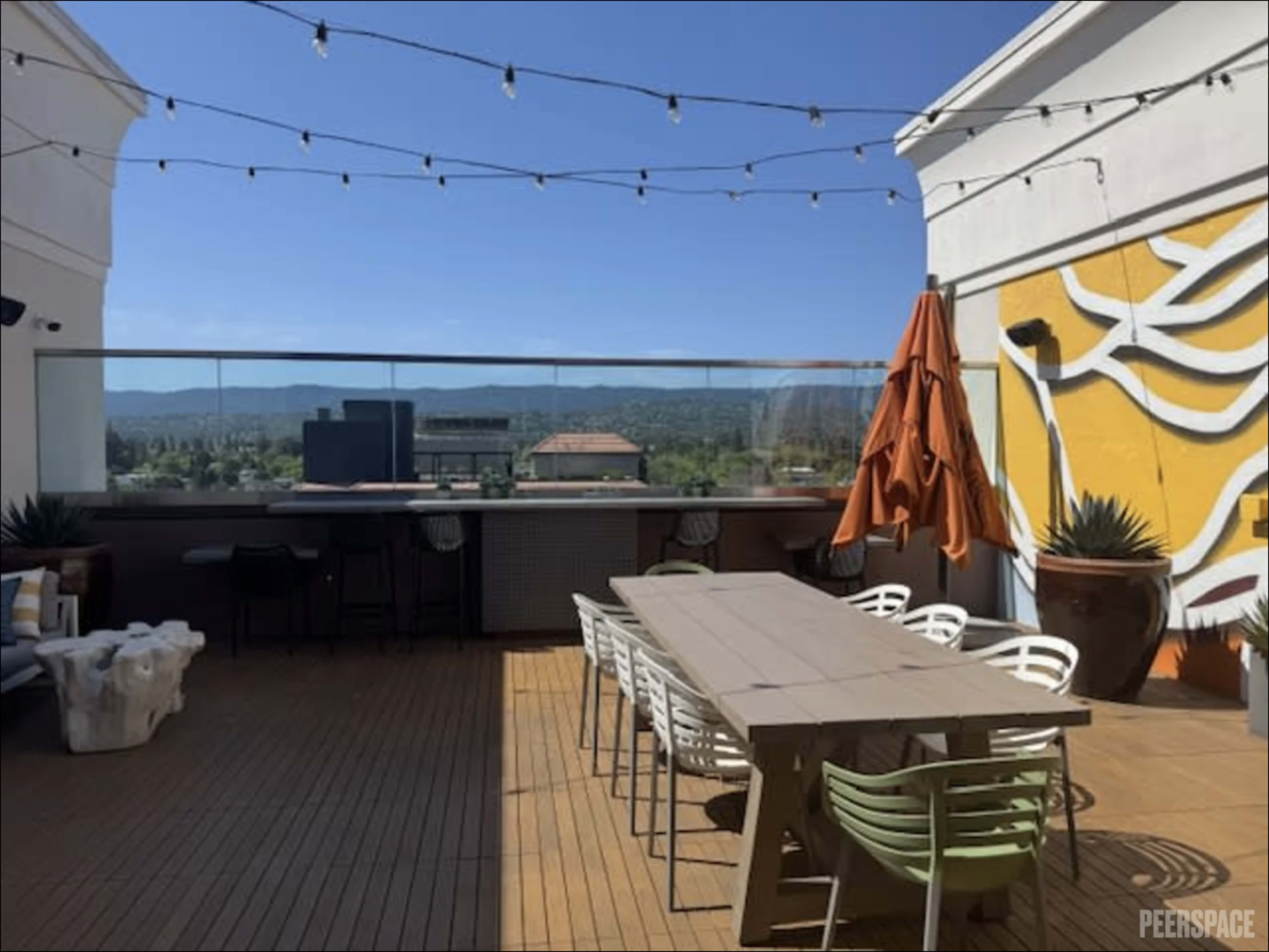 The image shows a rooftop terrace with a wooden deck, a long table, string lights, an orange umbrella, and a mural on the wall against a clear blue sky.