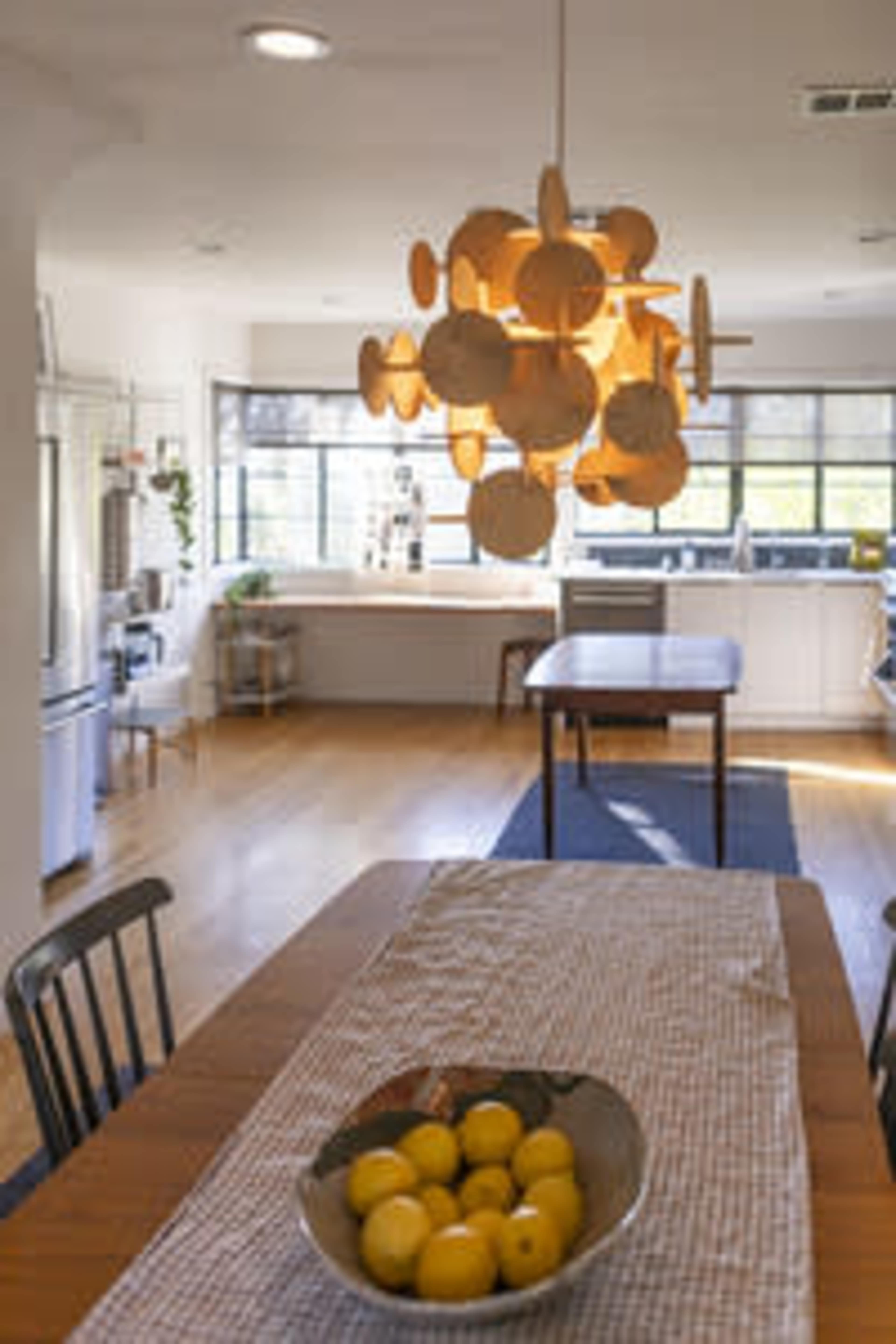 The image shows a modern kitchen with a wooden dining table set with a bowl of lemons, a unique hanging light fixture, and large windows allowing natural light.
