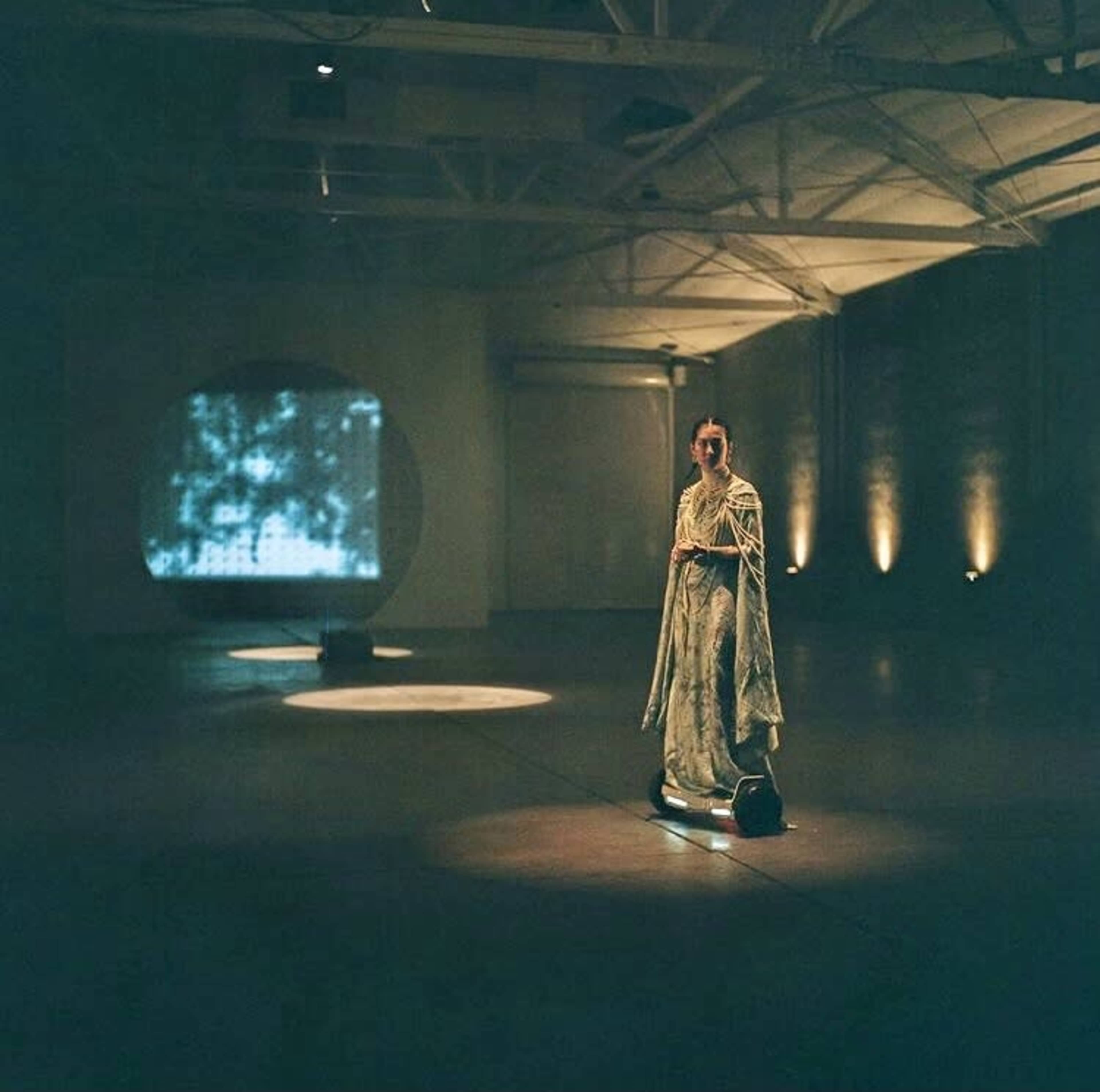 A woman in a flowing garment stands in an art gallery illuminated by projections on the walls and floor.