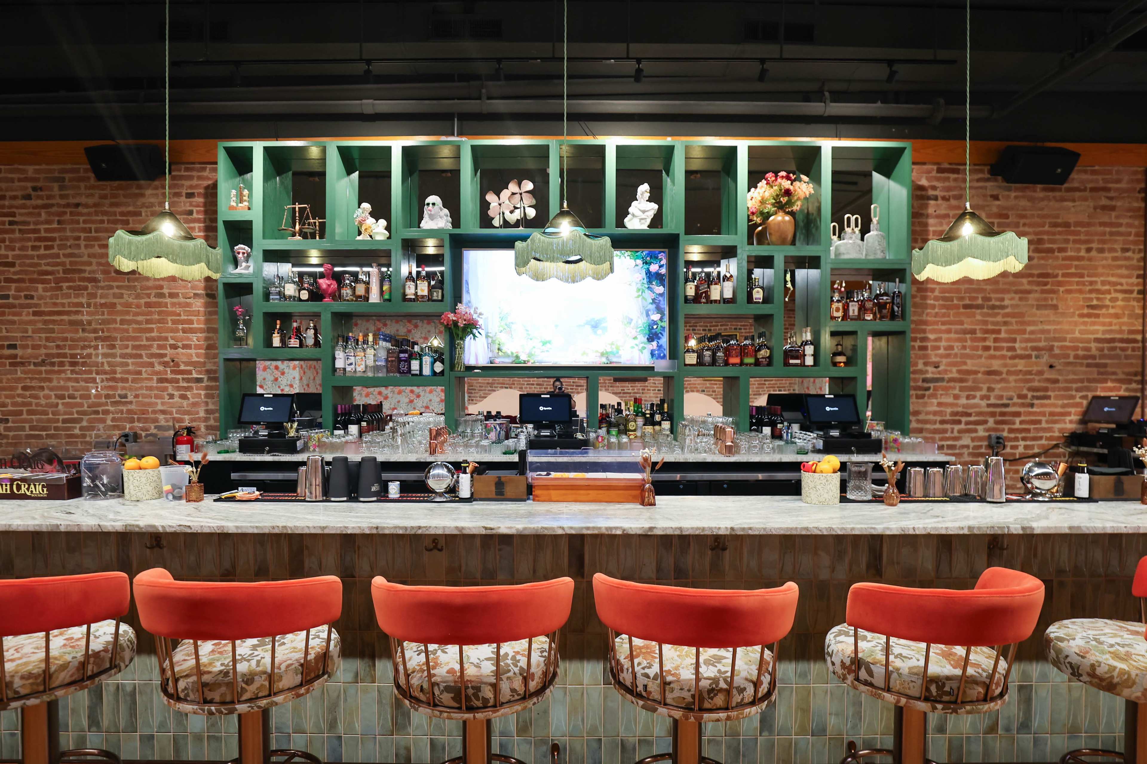A bar with a wooden counter and tall stools faces a green shelf display filled with bottles, decorative items, and a screen, against a backdrop of exposed brick walls.