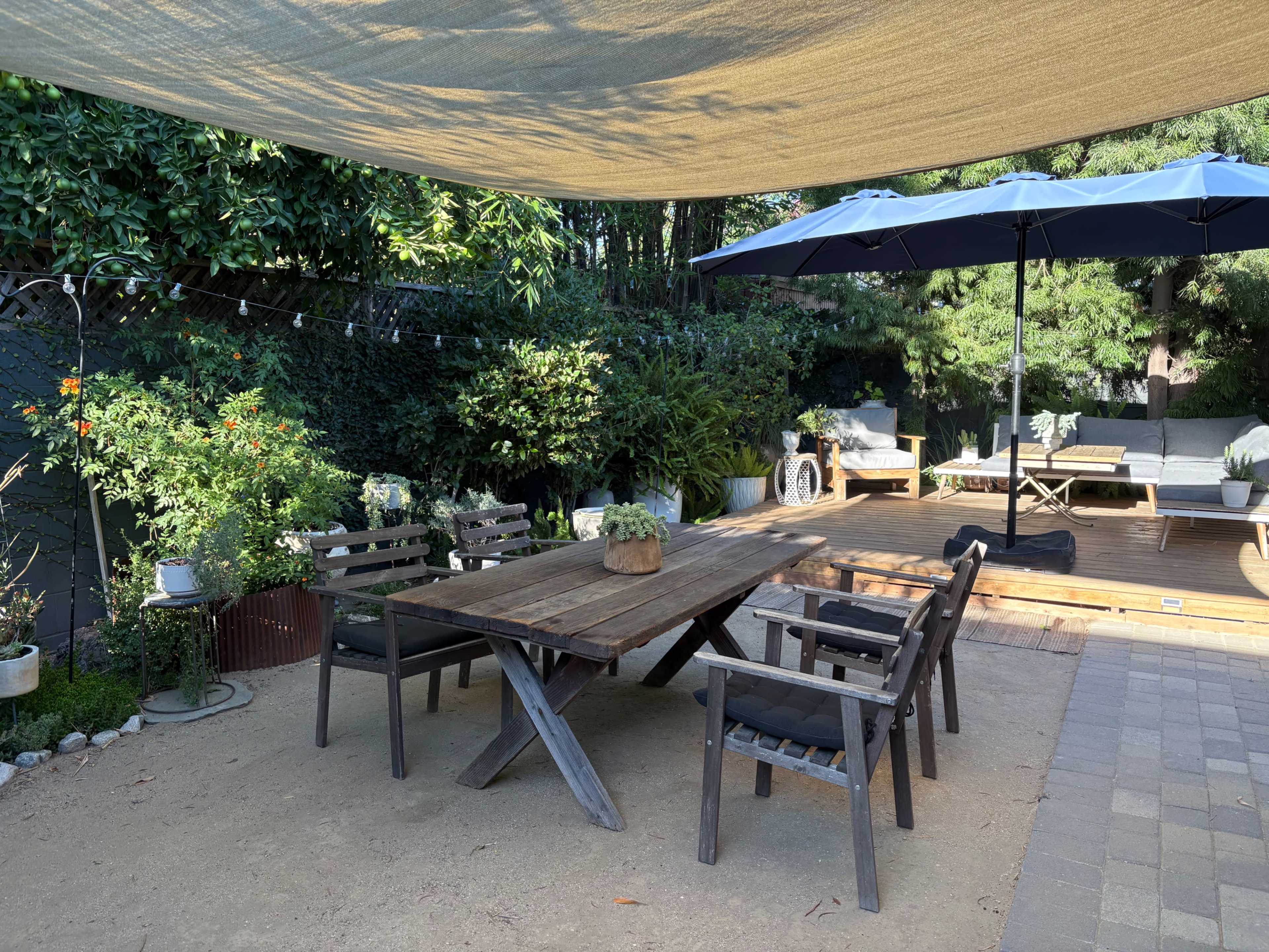 The image shows a shaded outdoor dining area with a wooden table surrounded by chairs, set against a backdrop of greenery and a deck.