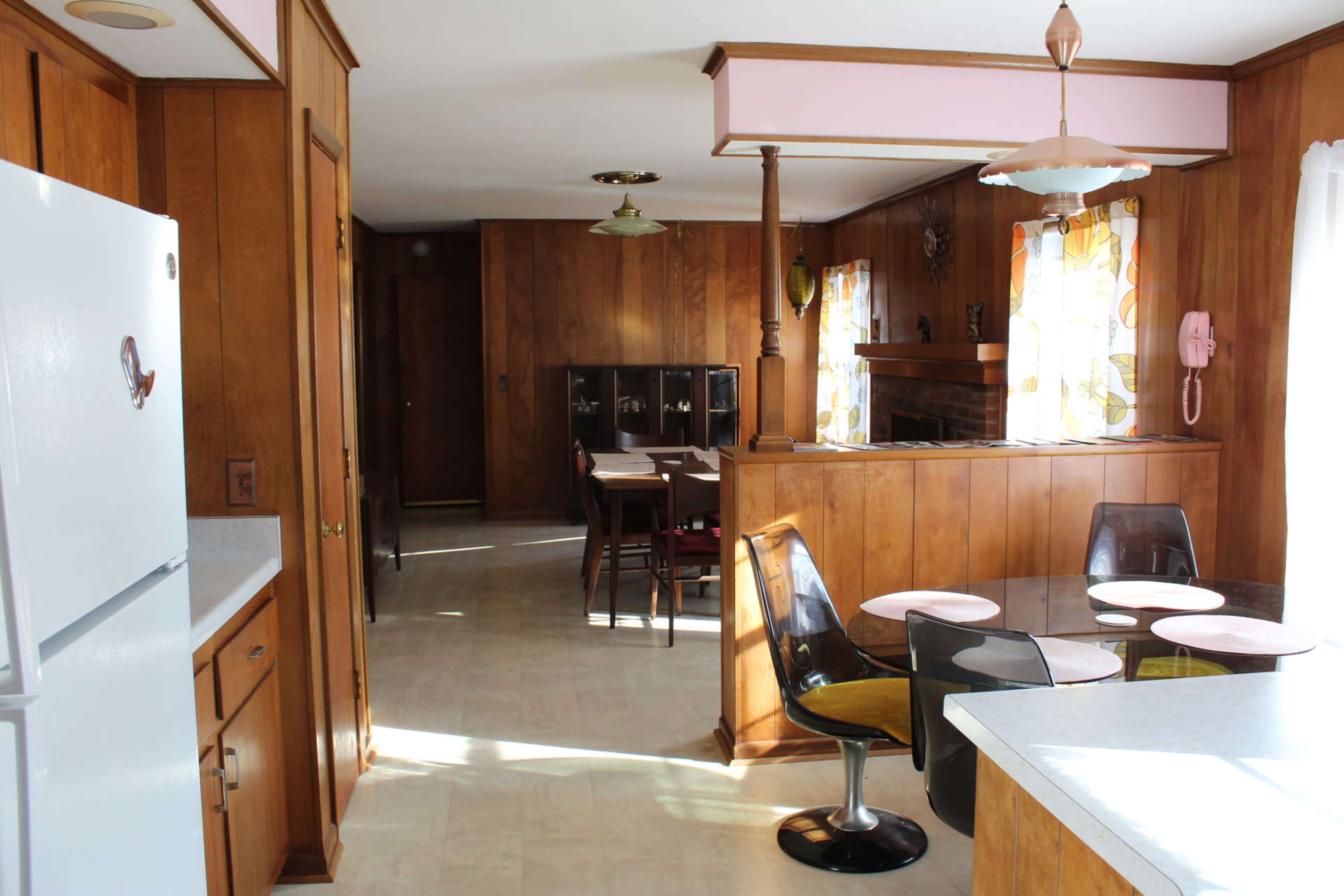 The image shows a spacious kitchen with wooden paneling, featuring a round dining table with chairs and an adjacent dining area with a large window adorned with colorful curtains.
