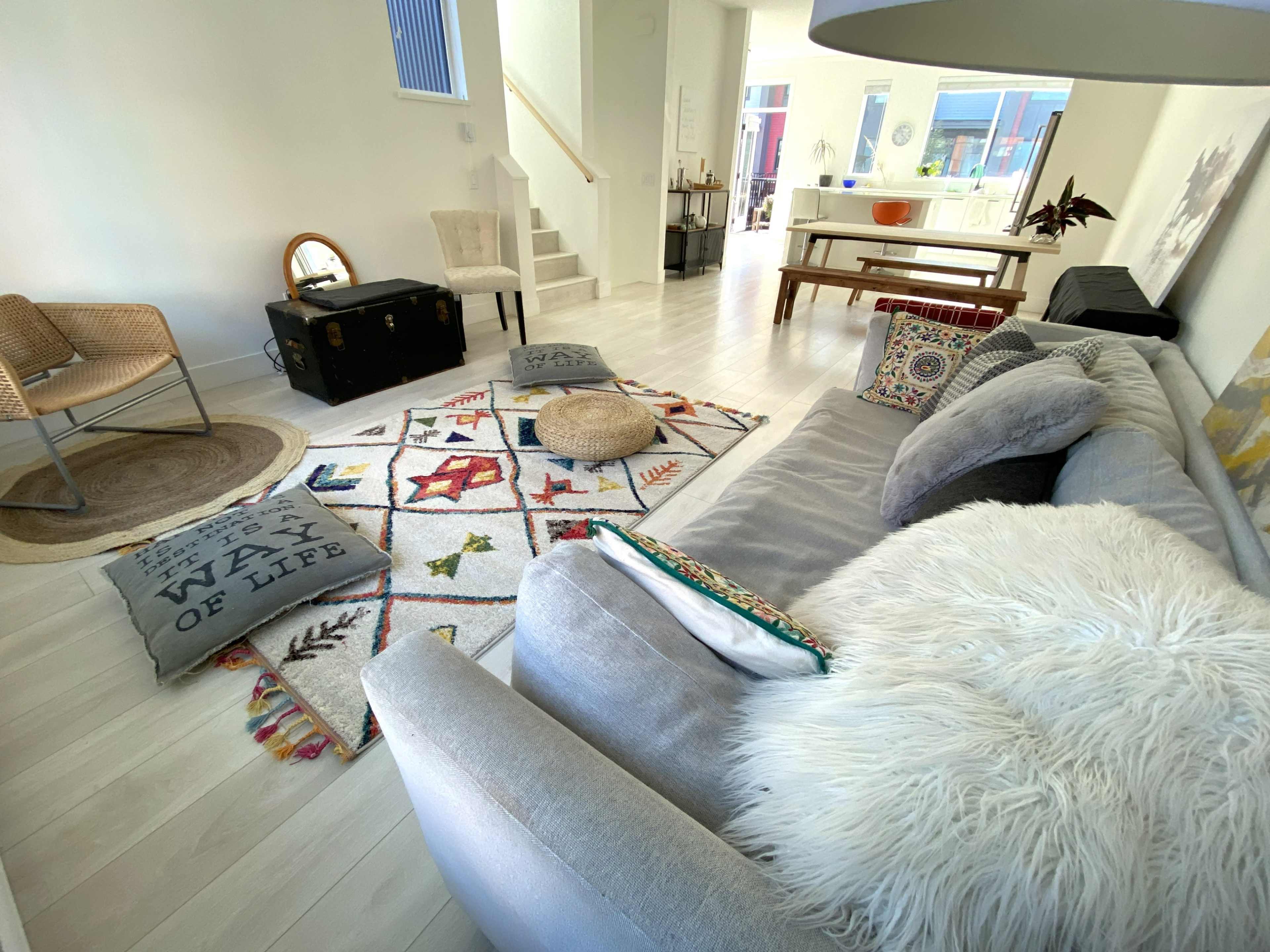 A bright living room with a gray sofa, a patterned area rug, a wooden dining table, and various decorative pillows and items.