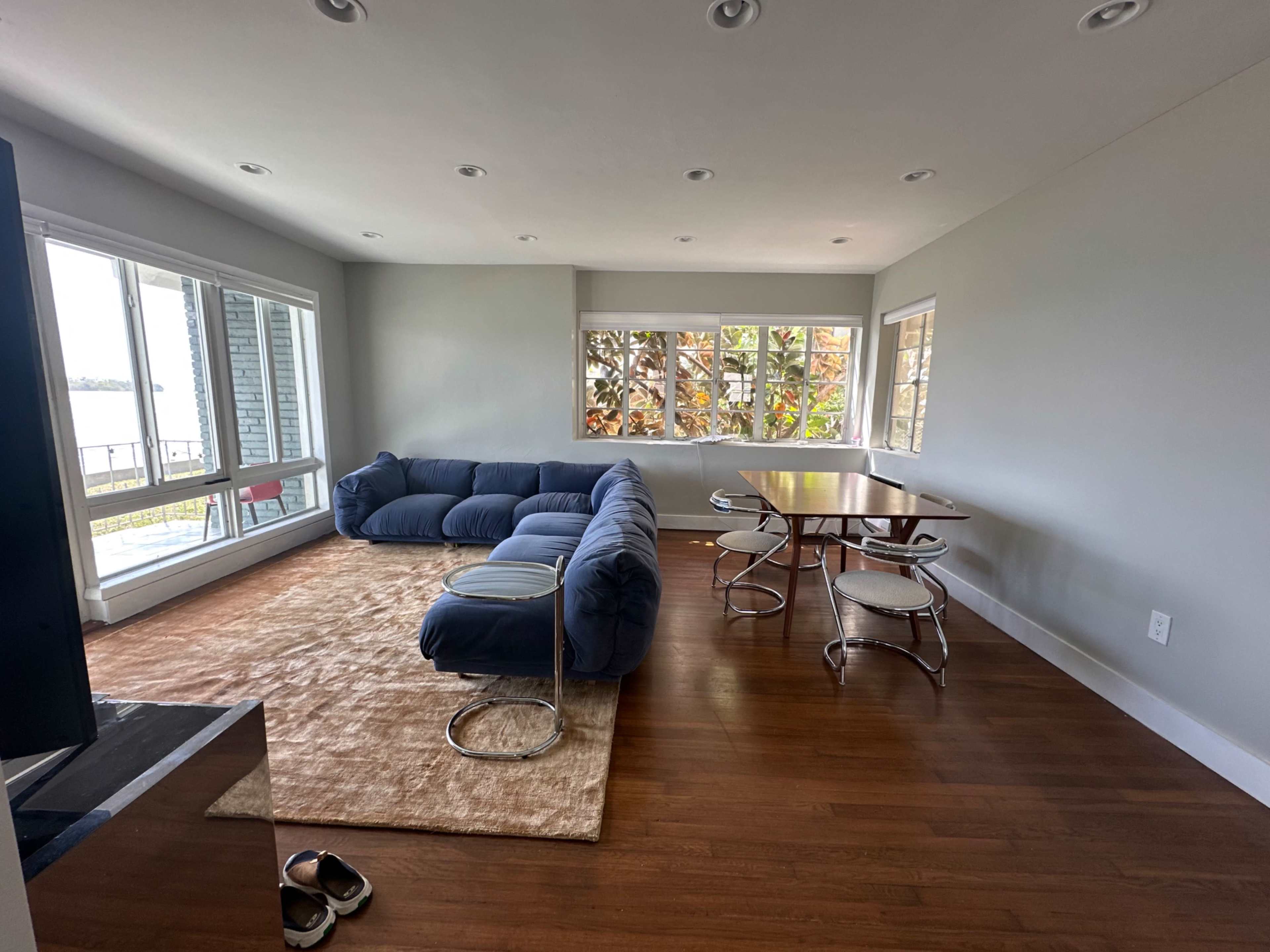 The image shows a modern living room with a blue sectional couch, a small table with chairs, and large windows allowing natural light to enter.