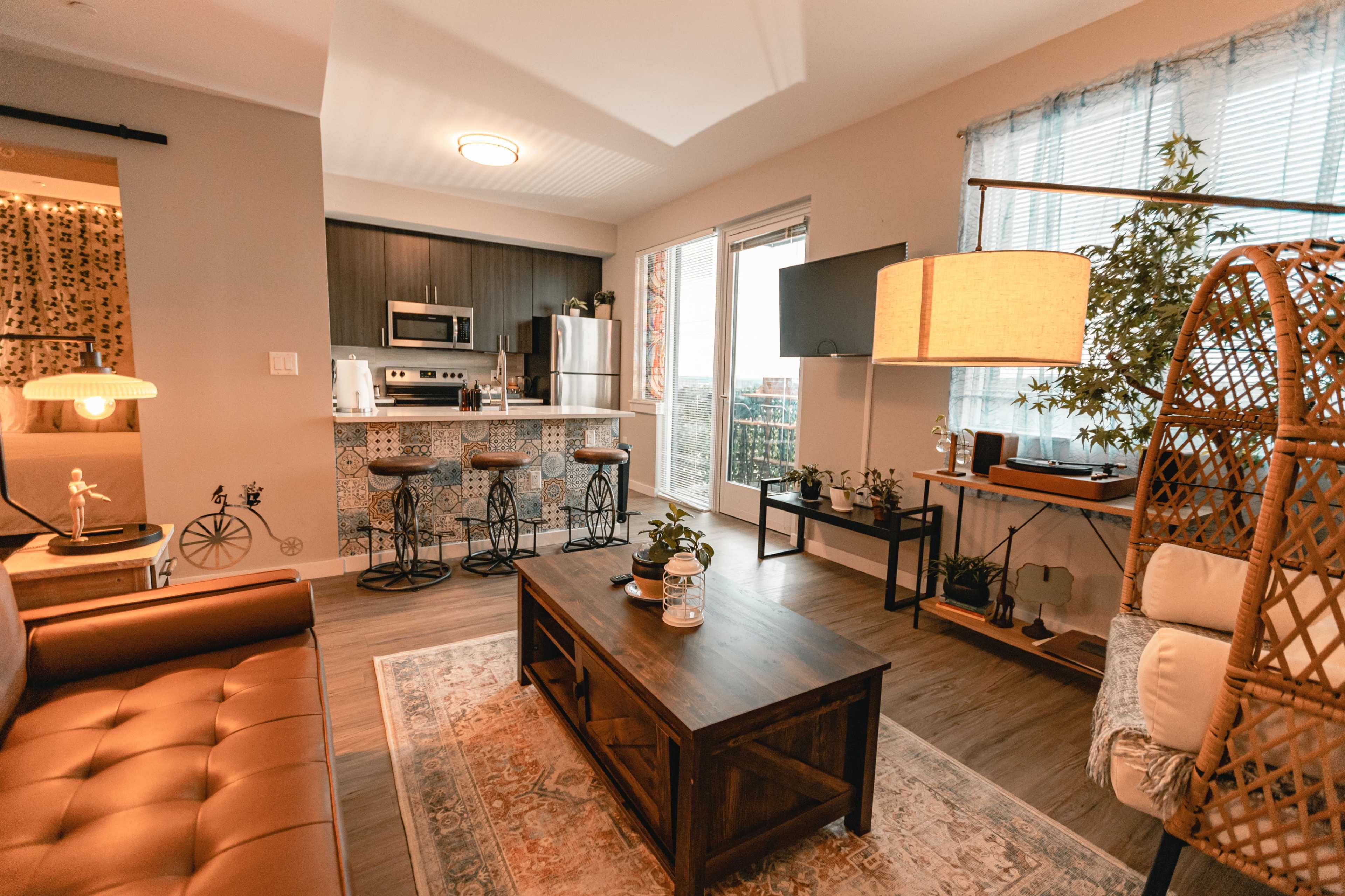 A modern living room with a brown leather couch, a wooden coffee table, a kitchen in the background featuring bar stools, and large windows with natural light.