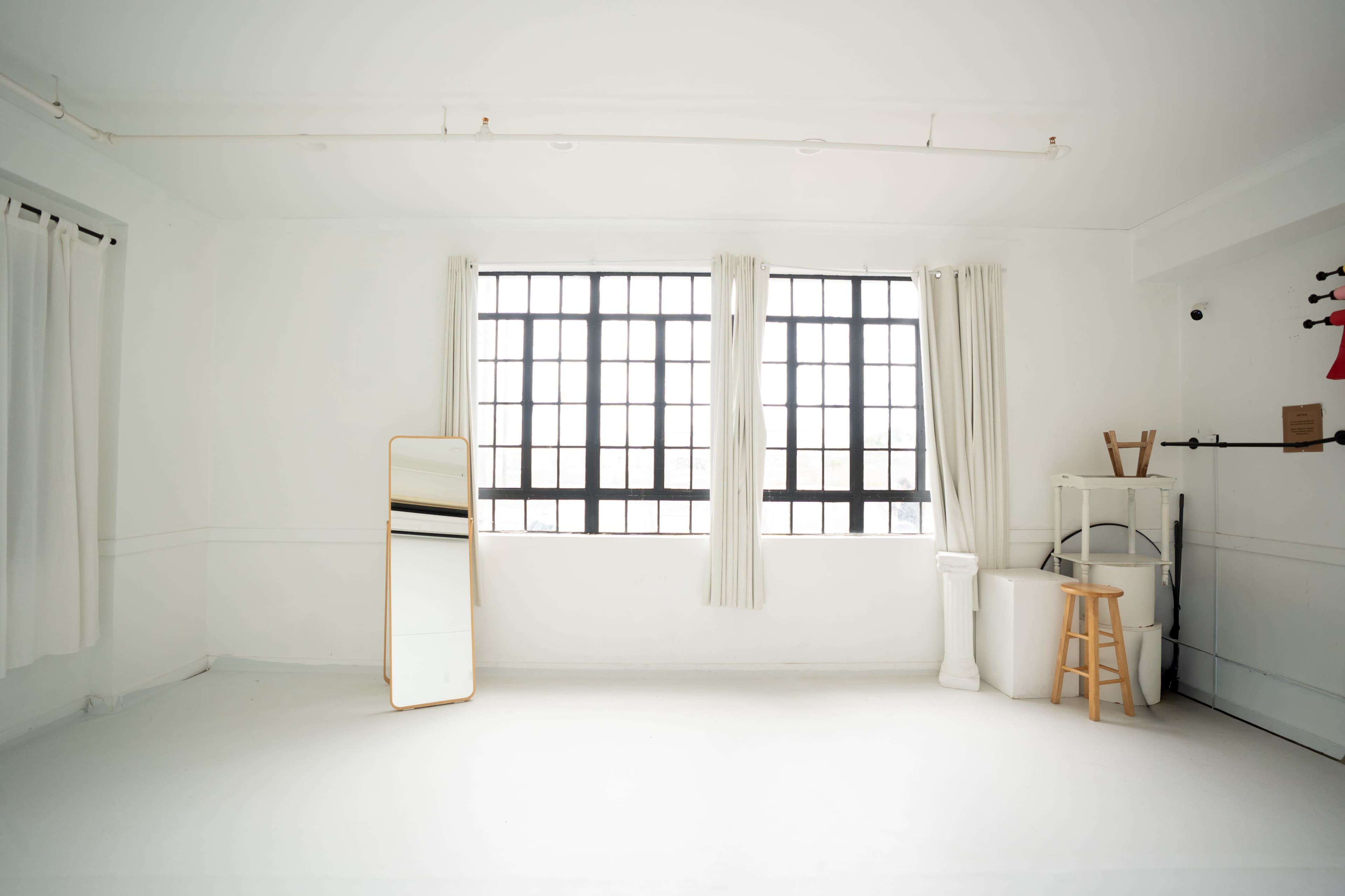 The image shows a bright, minimalist room with large windows, white walls, and a mirror positioned against one wall.