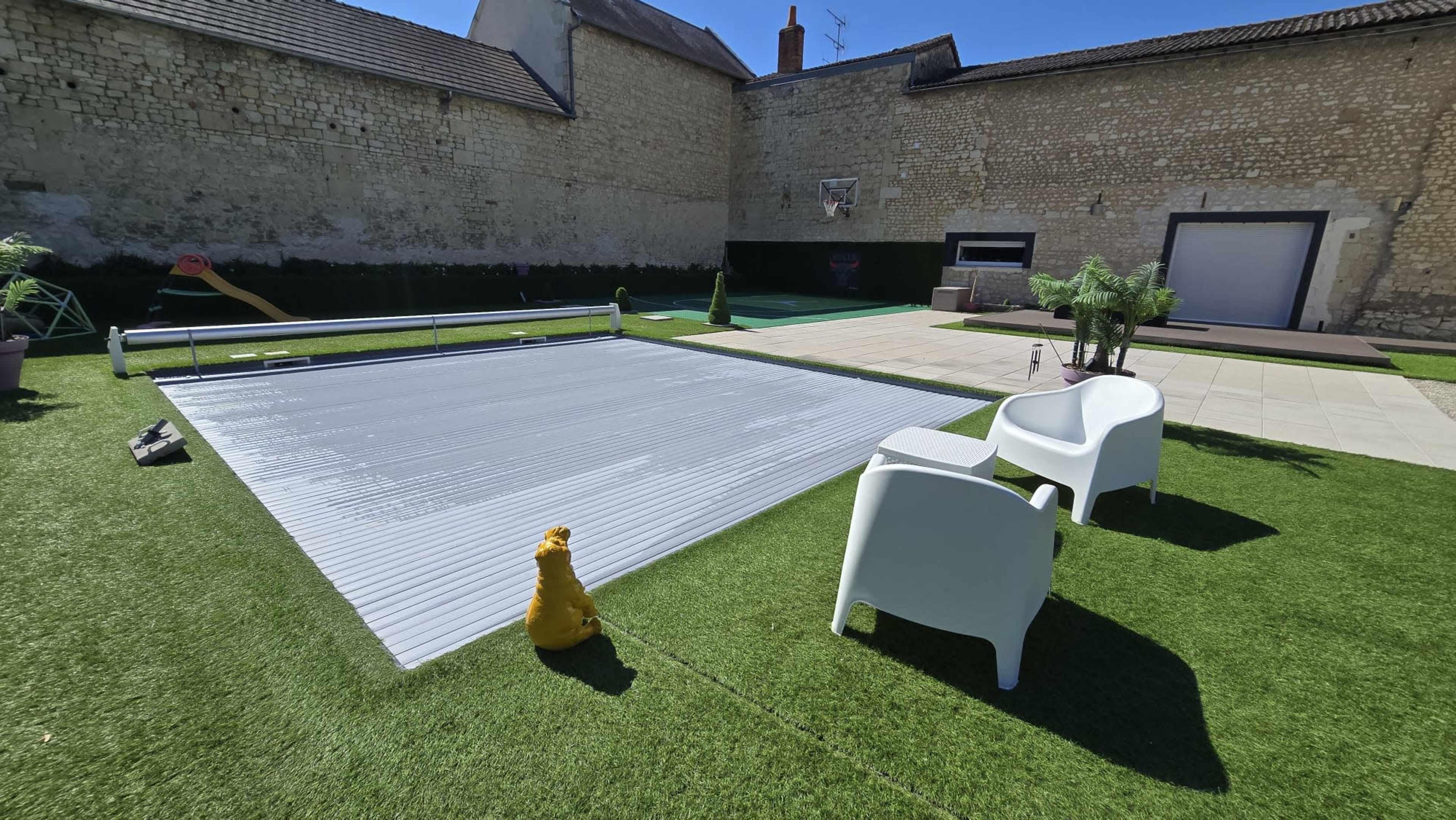 The image shows a sunny outdoor space featuring a covered swimming pool, two white chairs, and artificial grass, surrounded by stone walls and decorative plants.