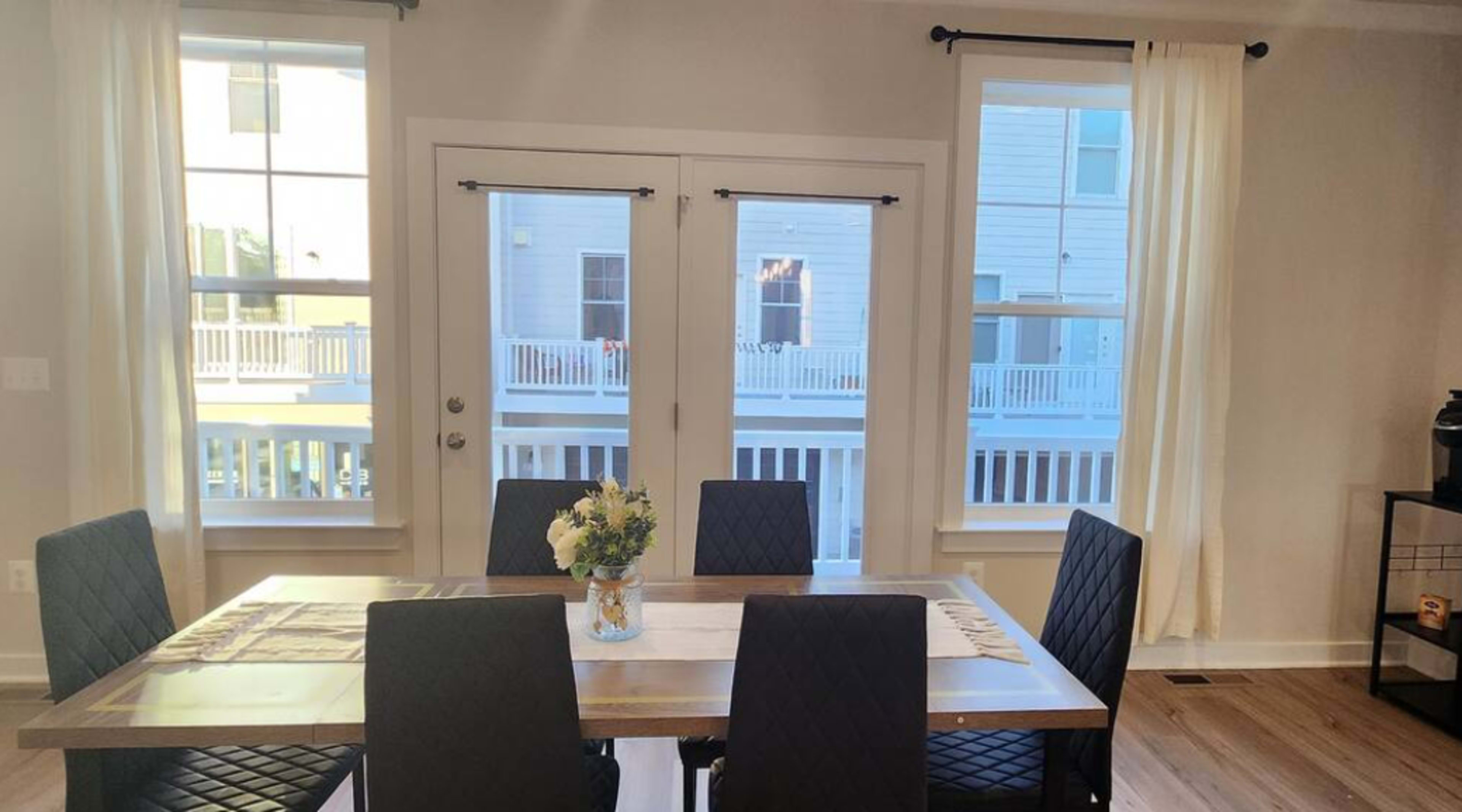 A dining table with six black chairs is positioned in a well-lit room featuring large windows and white curtains, overlooking a balcony area.