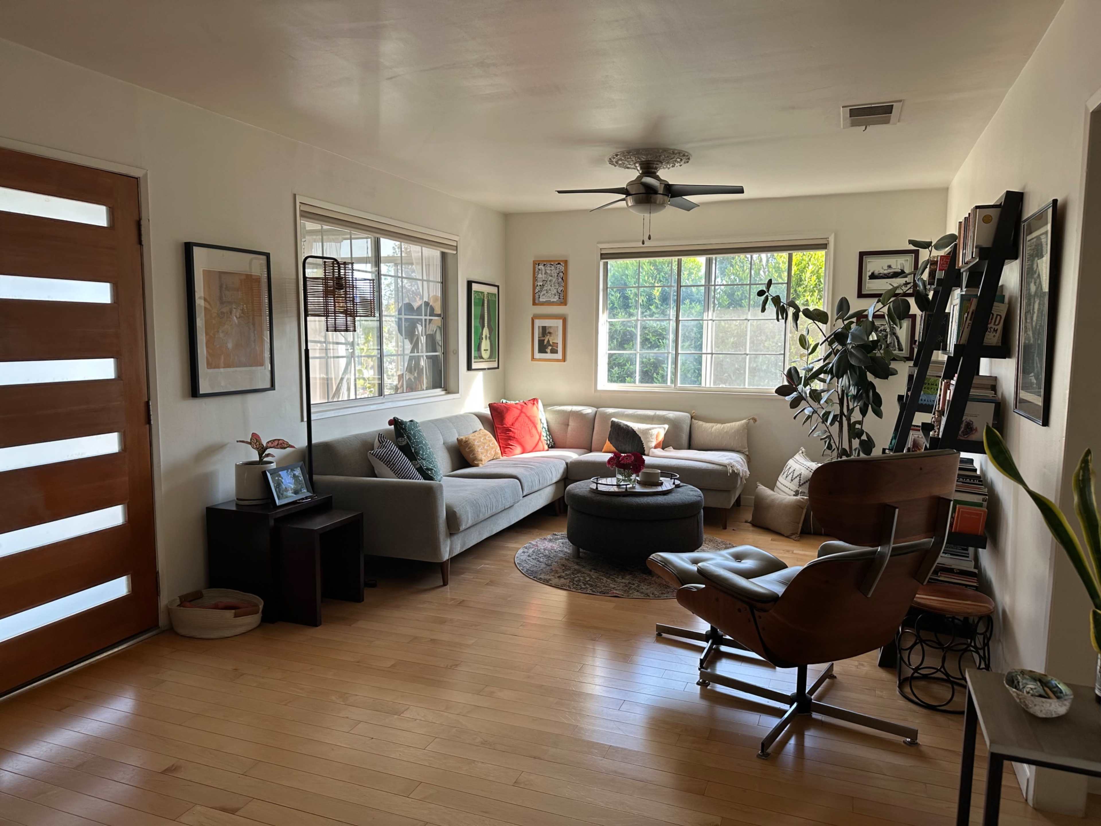 The image shows a living room with a gray sofa, a round coffee table, a wooden chair, and large windows allowing natural light to enter.