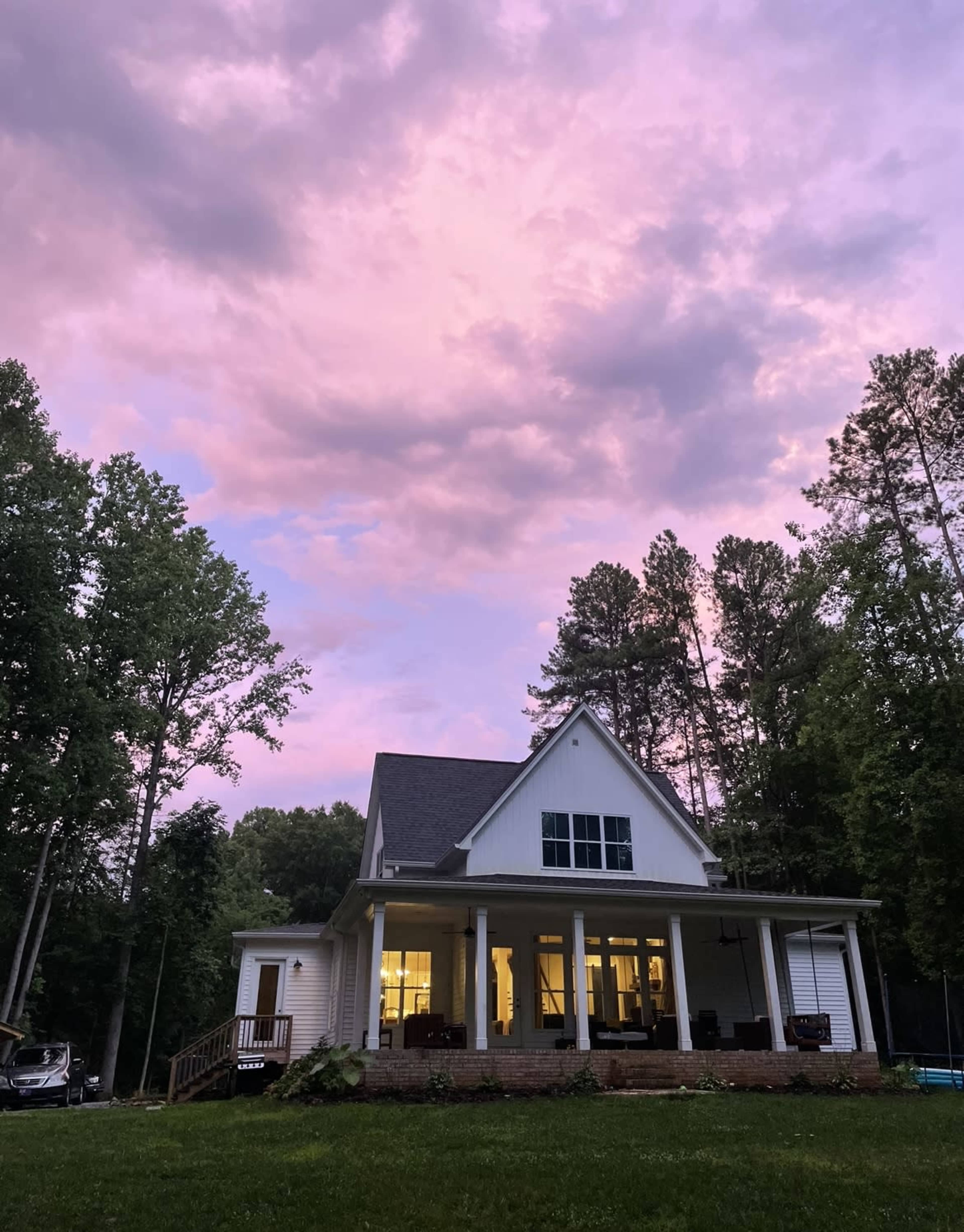 A white house with a porch sits in a forested area under a pink and purple sky at dusk.