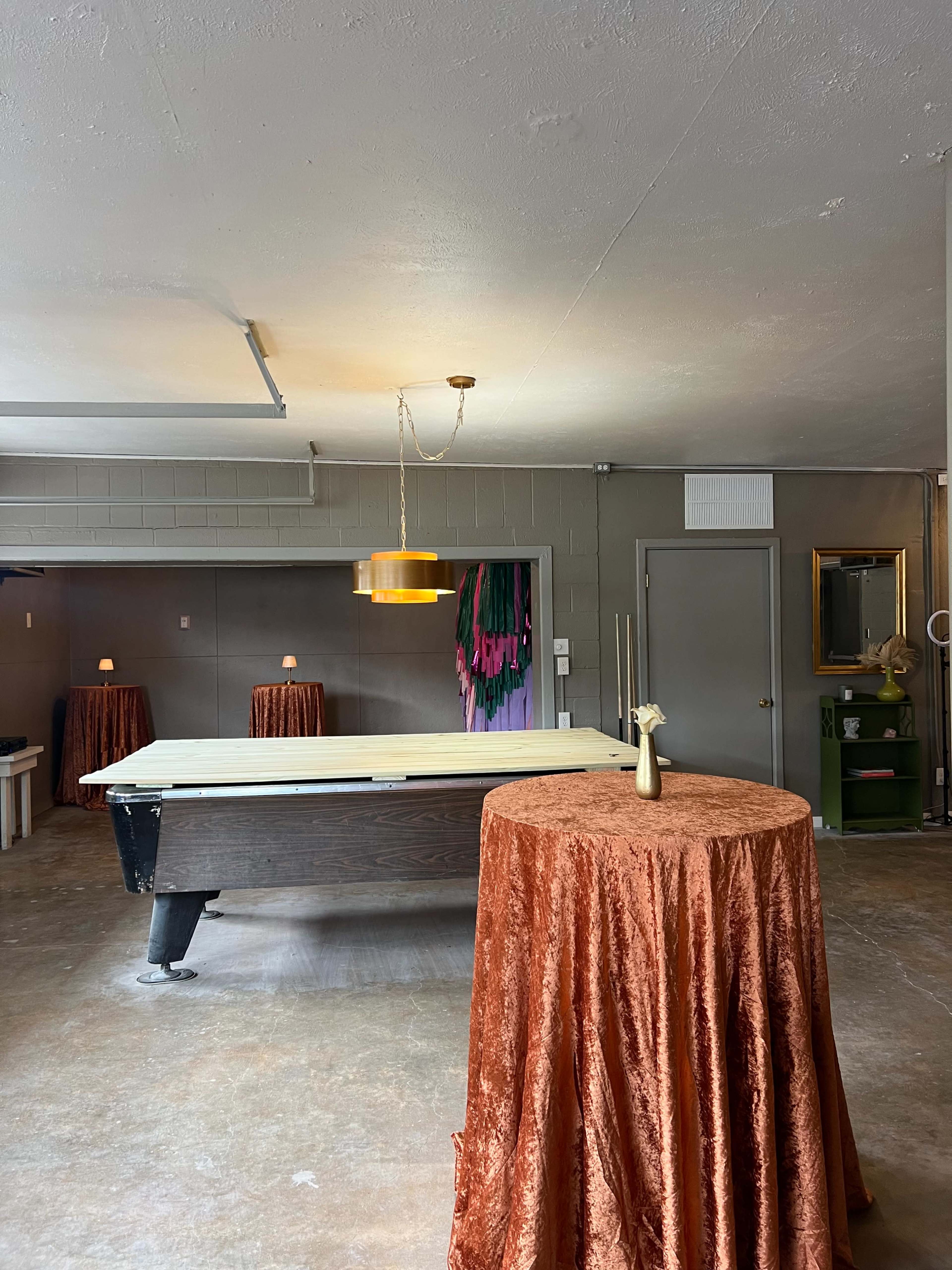 The image shows a spacious room with a pool table and two round tables covered in orange tablecloths, along with a hanging light fixture above.