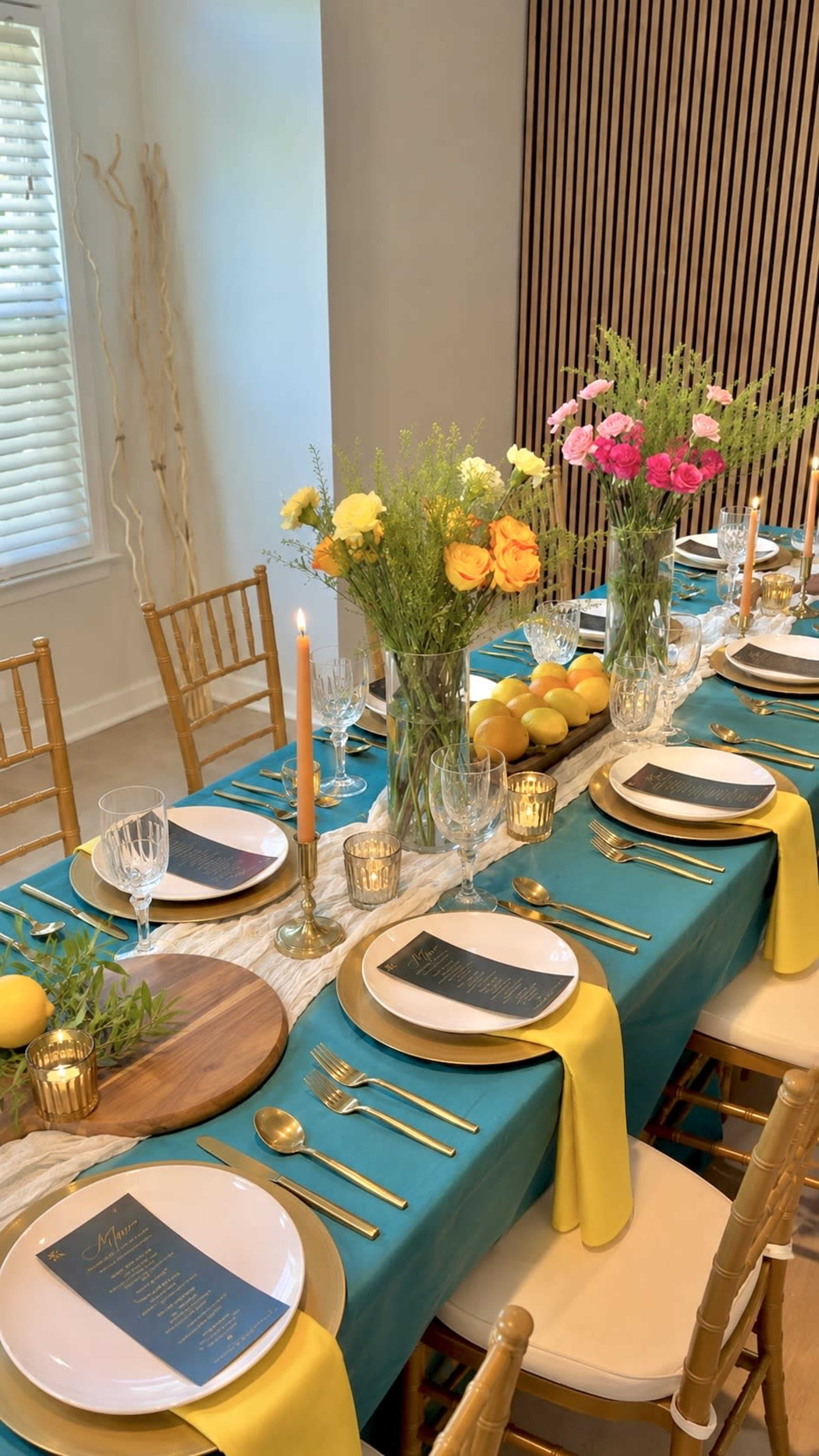 A dining table is elegantly set with a turquoise tablecloth, gold utensils, white plates, and colorful floral centerpieces, surrounded by chairs with yellow sashes.