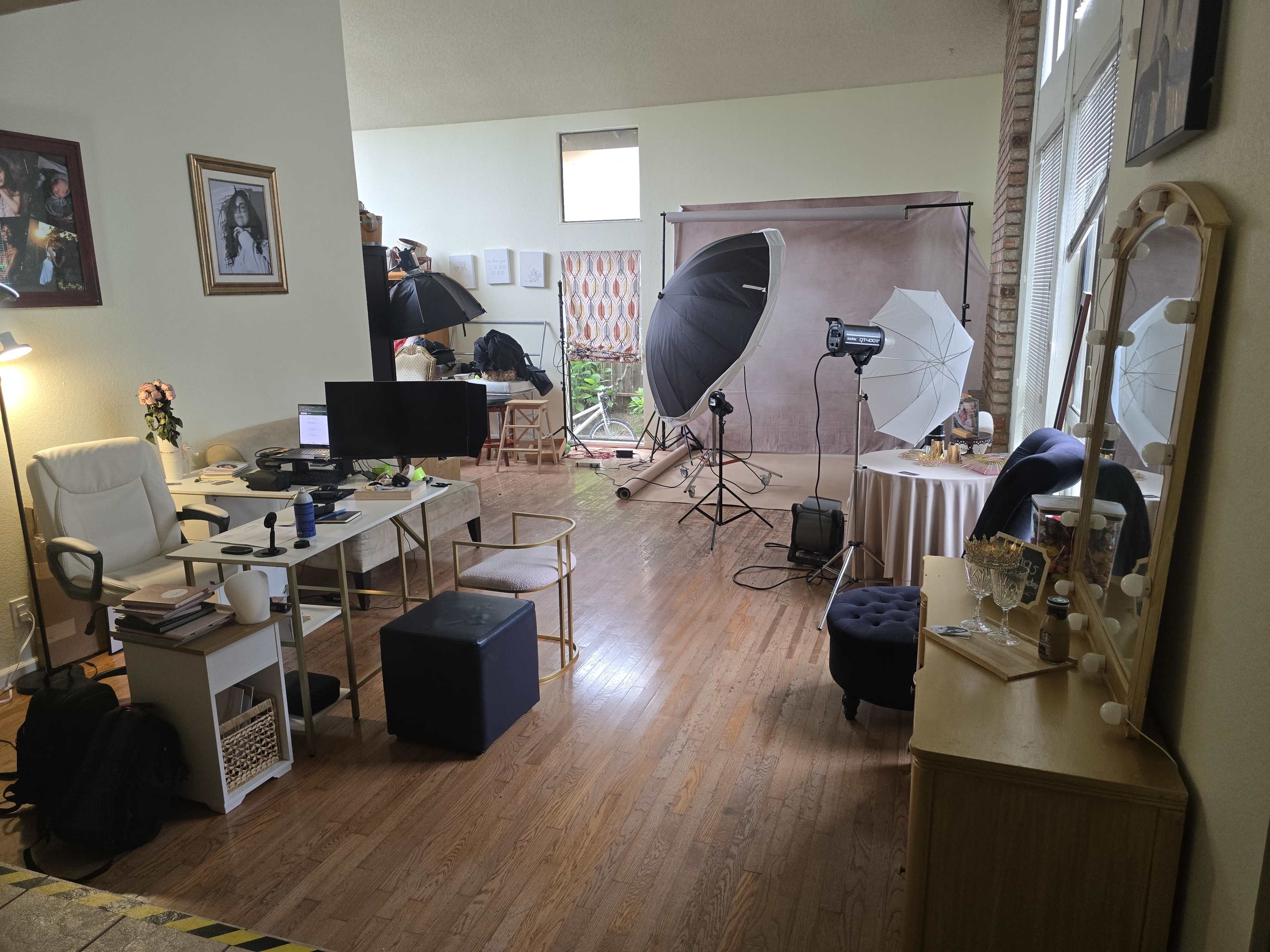 The image shows a well-lit photography studio featuring a desk with a computer, chairs, softboxes, and a backdrop set up for shoots.