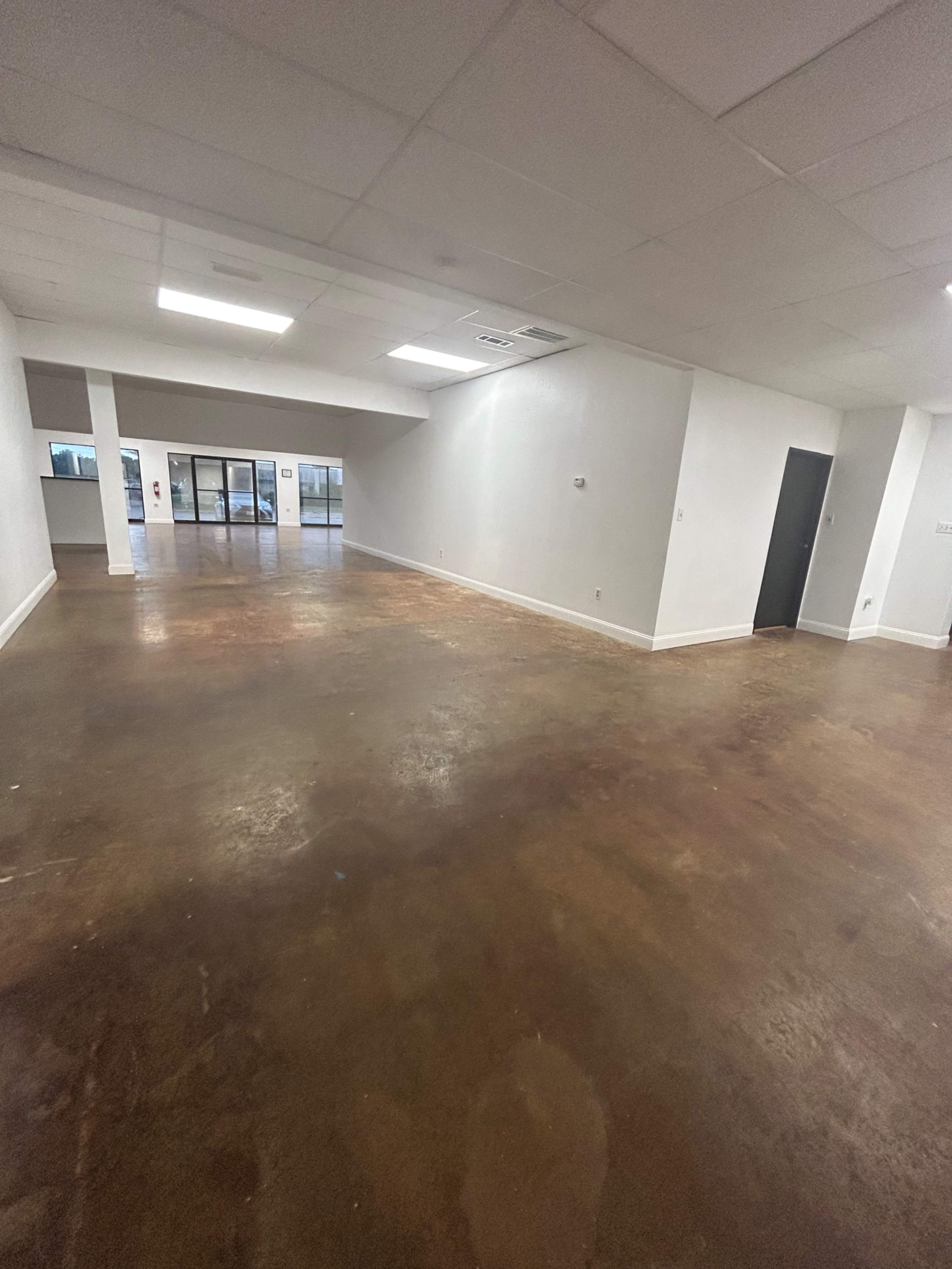 The image shows an empty room with polished, stained concrete flooring and white walls, featuring large windows and a black door on one side.