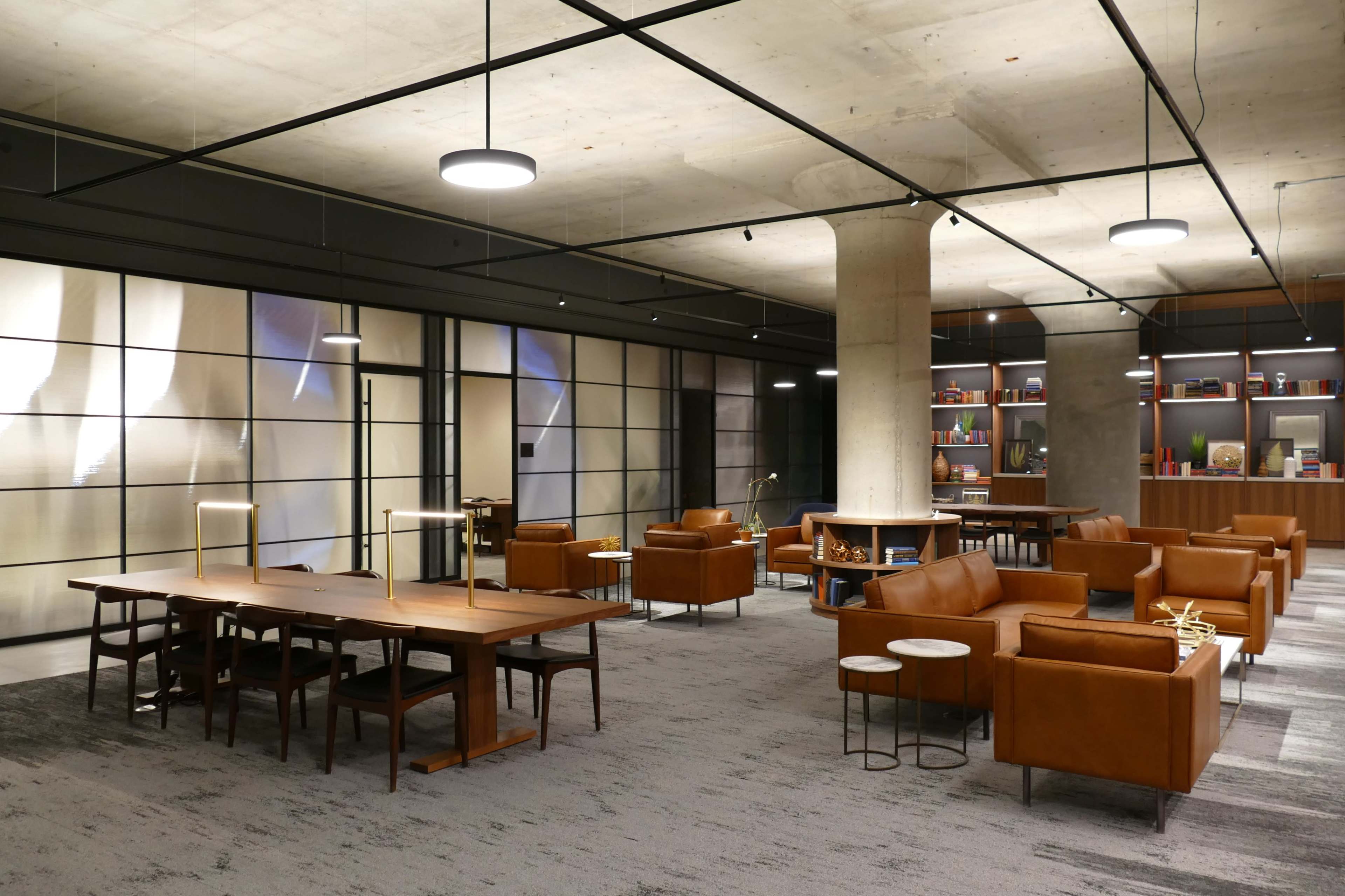 A modern office space featuring wooden tables and brown leather seating arranged in a well-lit area with exposed concrete pillars.