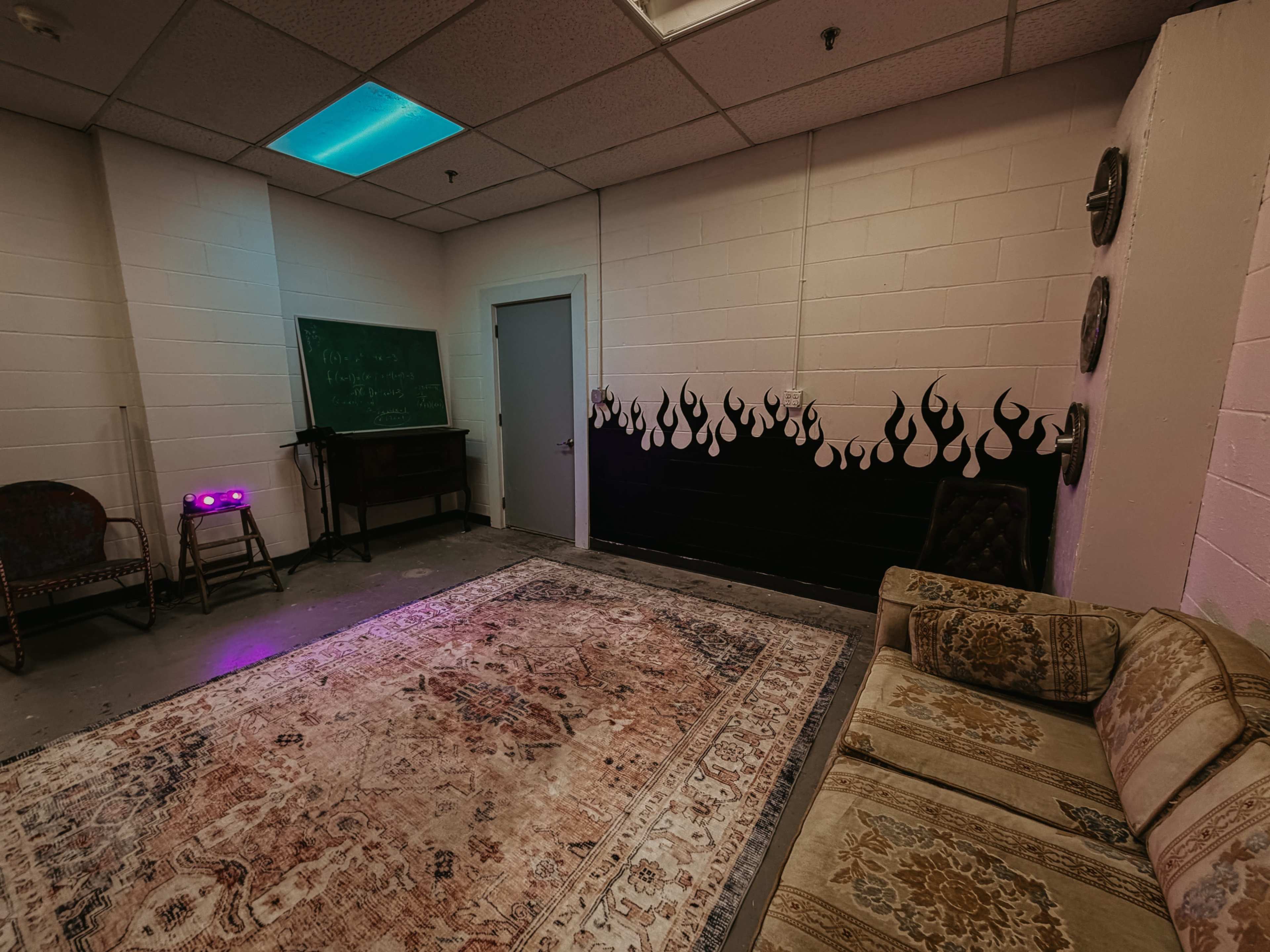 The image shows a small, dimly lit room with a large rug on the floor, a patterned couch, a chalkboard, and a wall painted with black flames.