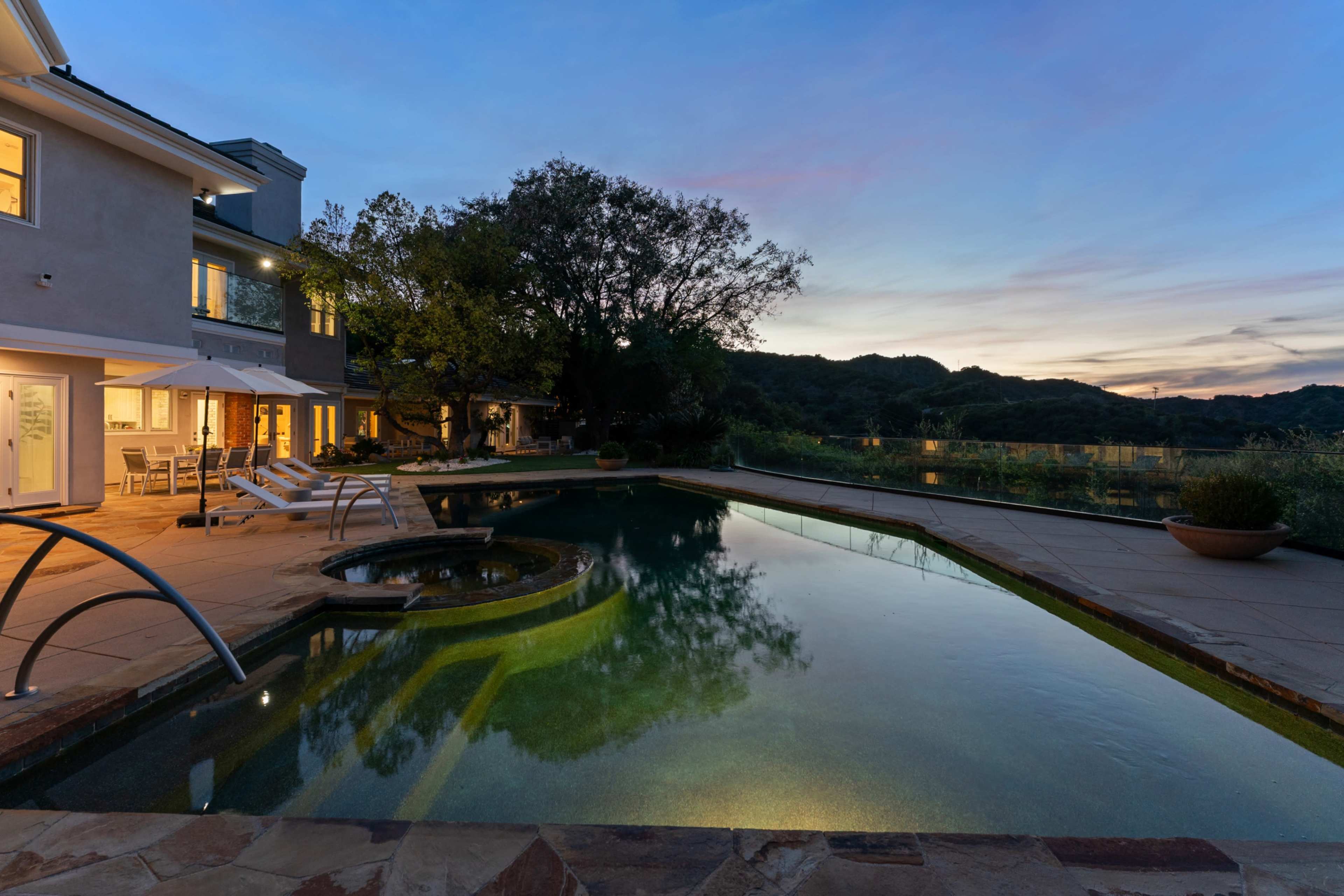 The image shows a serene backyard with a swimming pool and a jacuzzi, surrounded by trees and hills under a twilight sky.