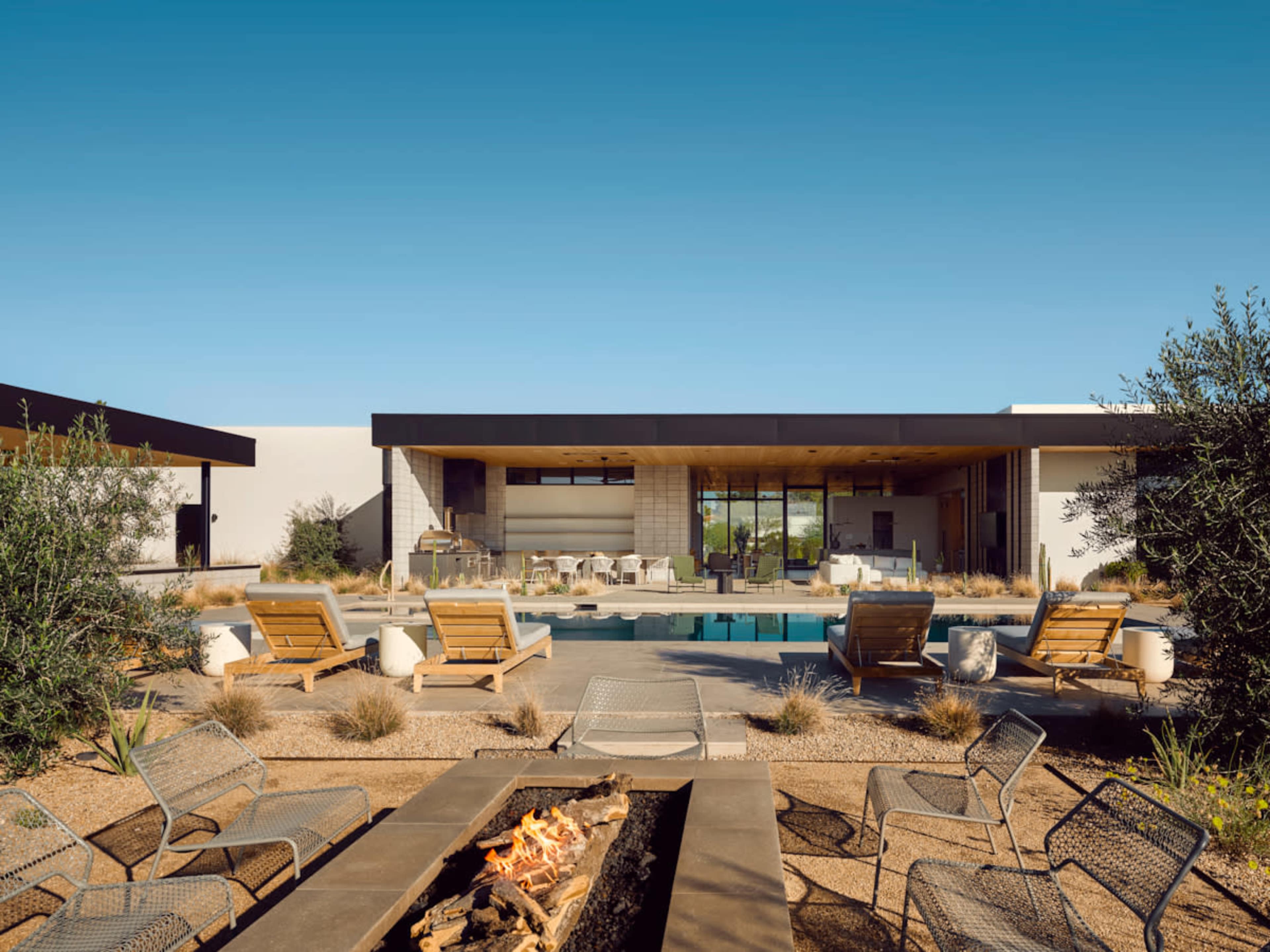 The image features a modern architectural home with a swimming pool surrounded by lounge chairs and a fire pit, set against a clear blue sky.