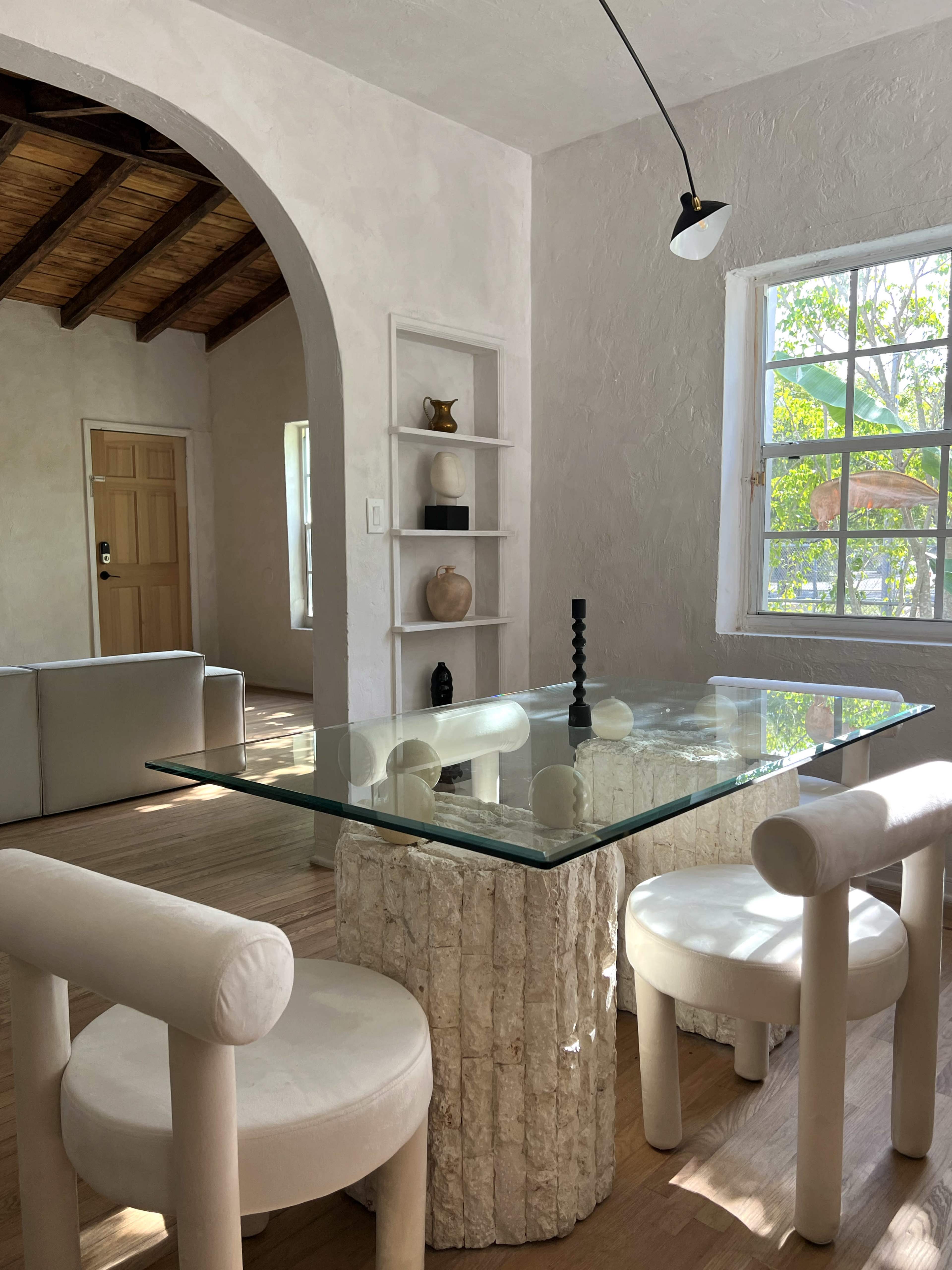 A dining area with a glass table supported by a textured stone base, surrounded by four tubular chairs, in a room with light-colored walls and wood accents.