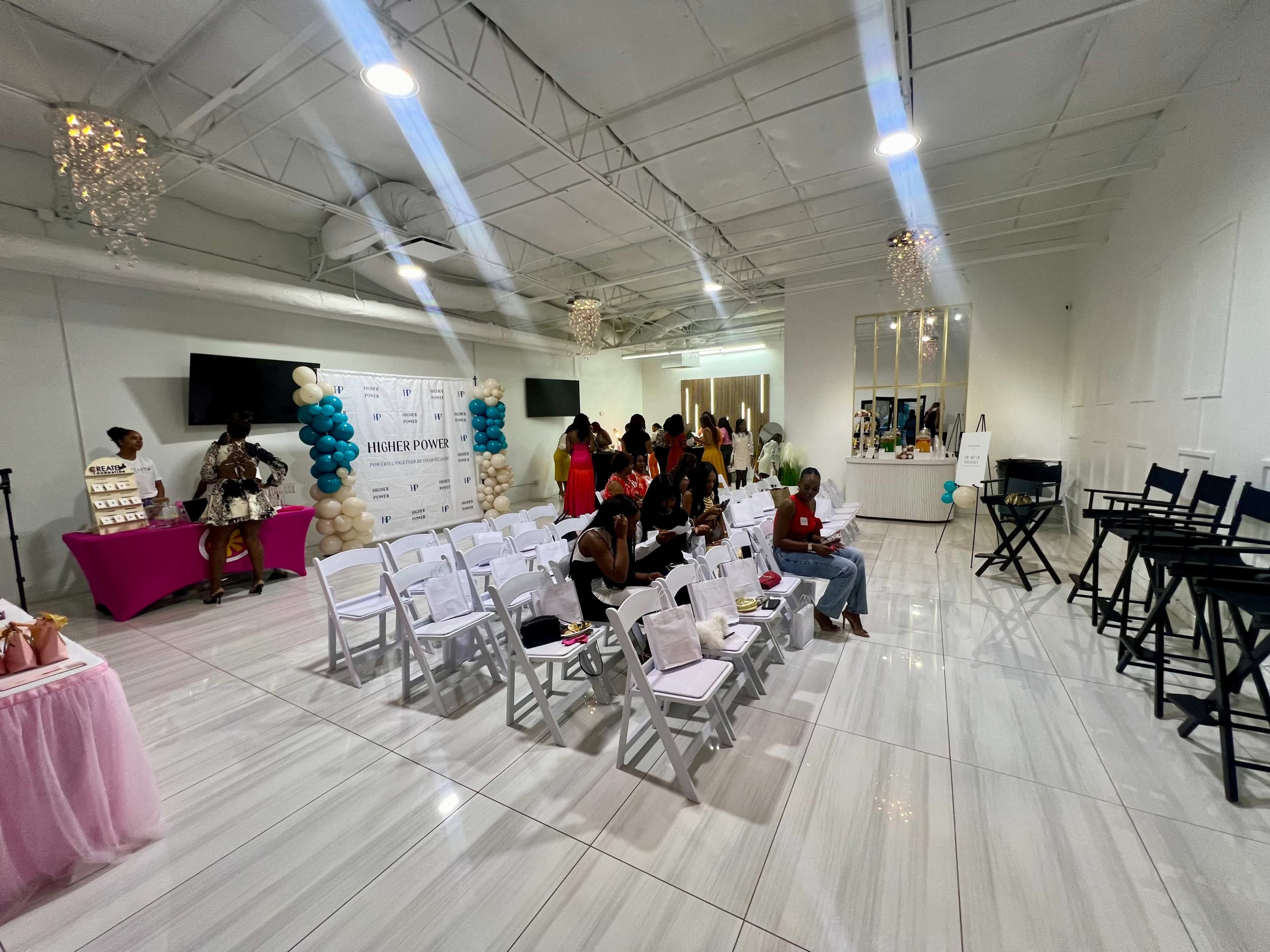 A spacious indoor venue is set up with rows of white chairs, a pink and purple table display, and guests mingling in the background.