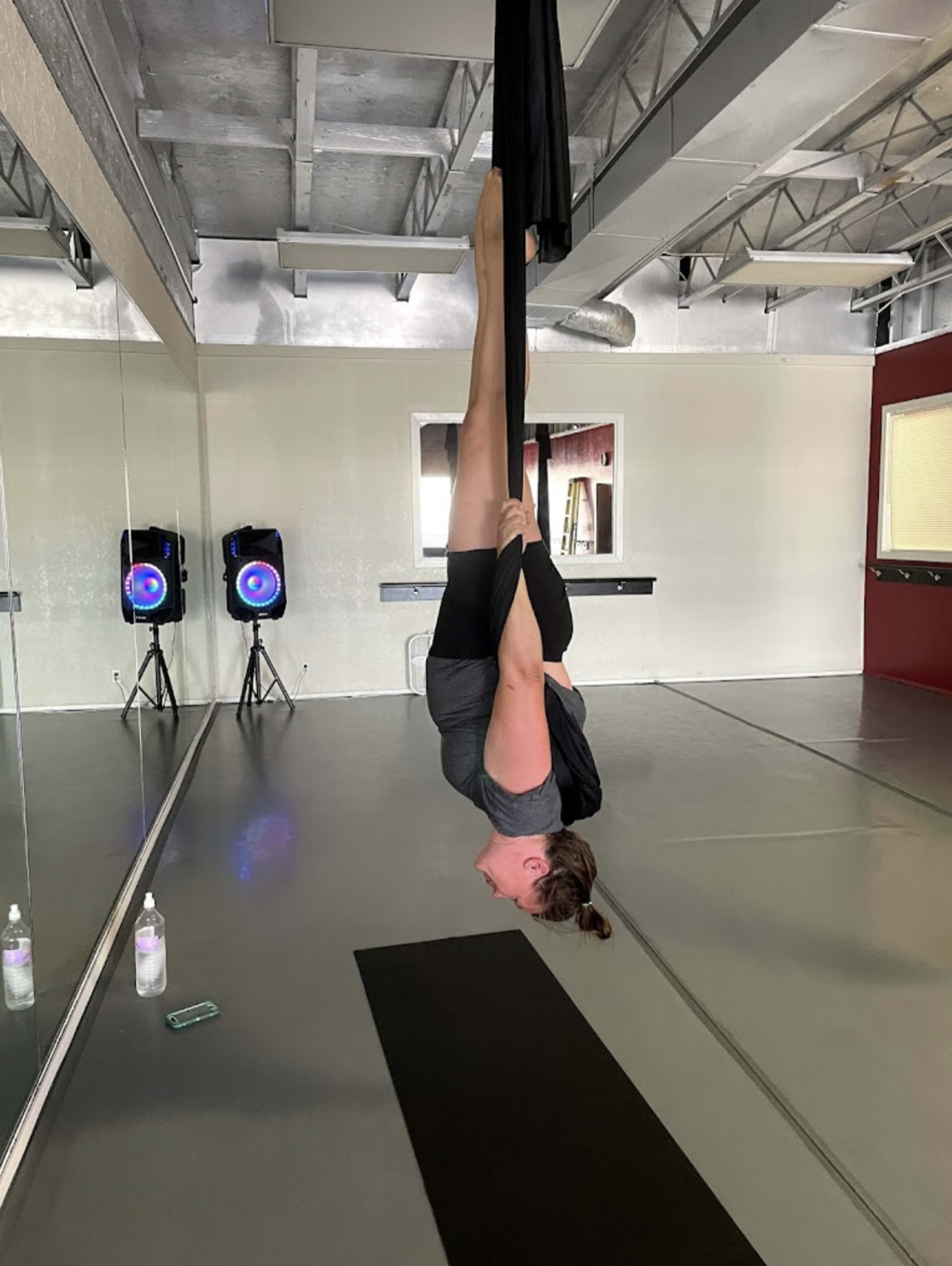 A person is performing an inverted pose using aerial silks in a spacious studio with mirrors and sound equipment in the background.