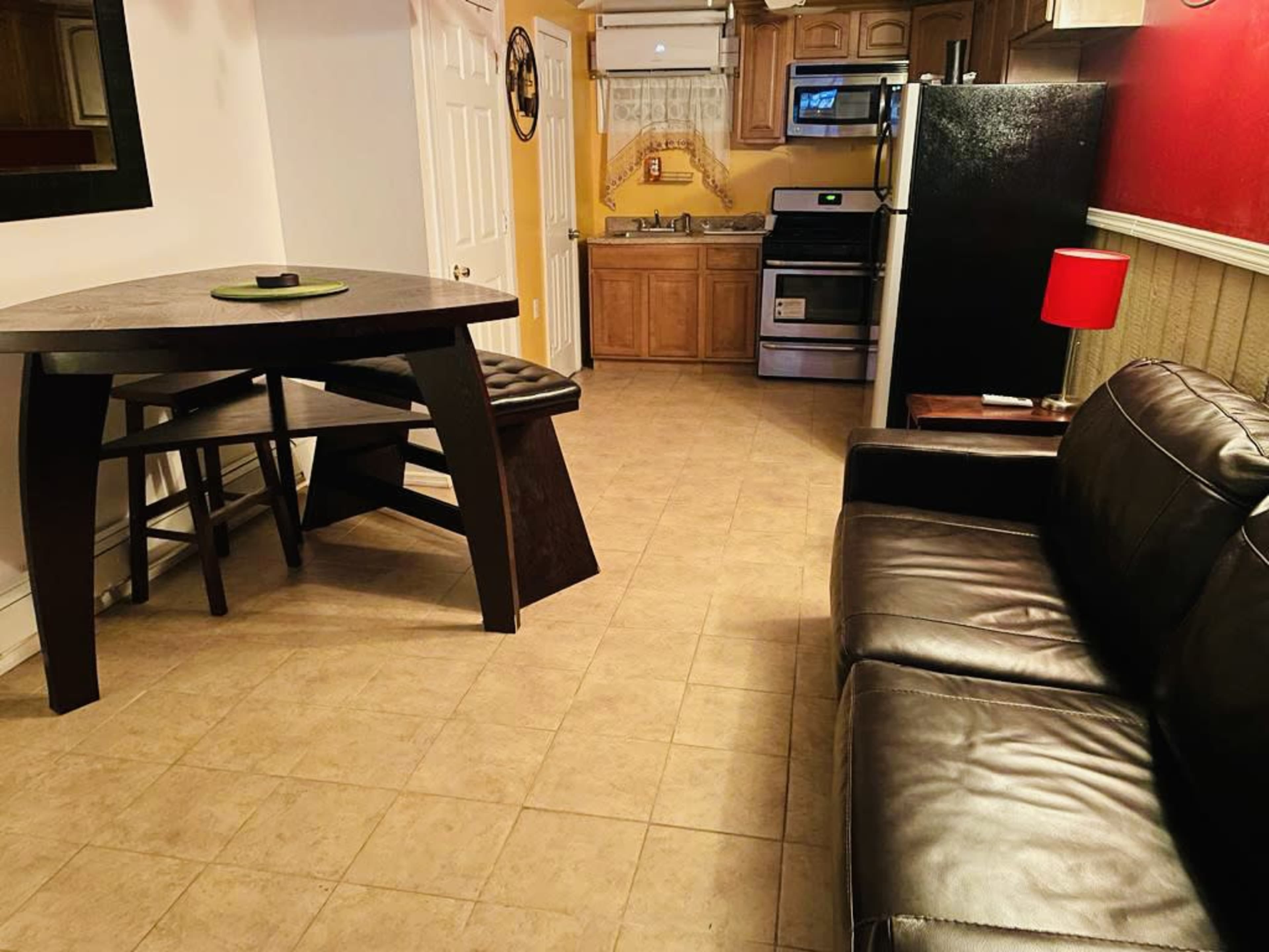 A small kitchen and living area featuring a black table with stools, a leather couch, and kitchen appliances, including a refrigerator and stove.