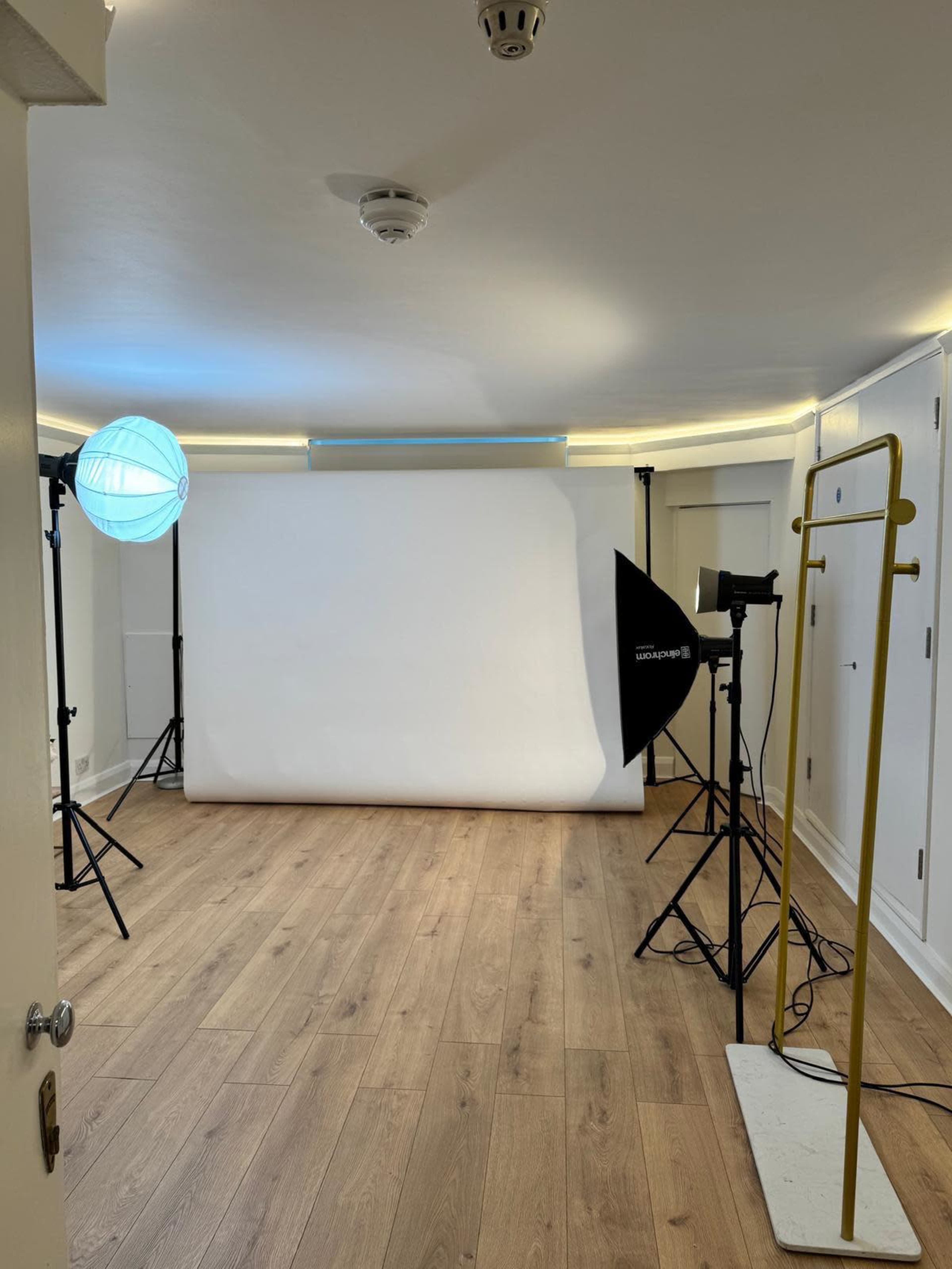 The image shows a photo studio setup with lighting equipment, a backdrop, and wooden flooring.