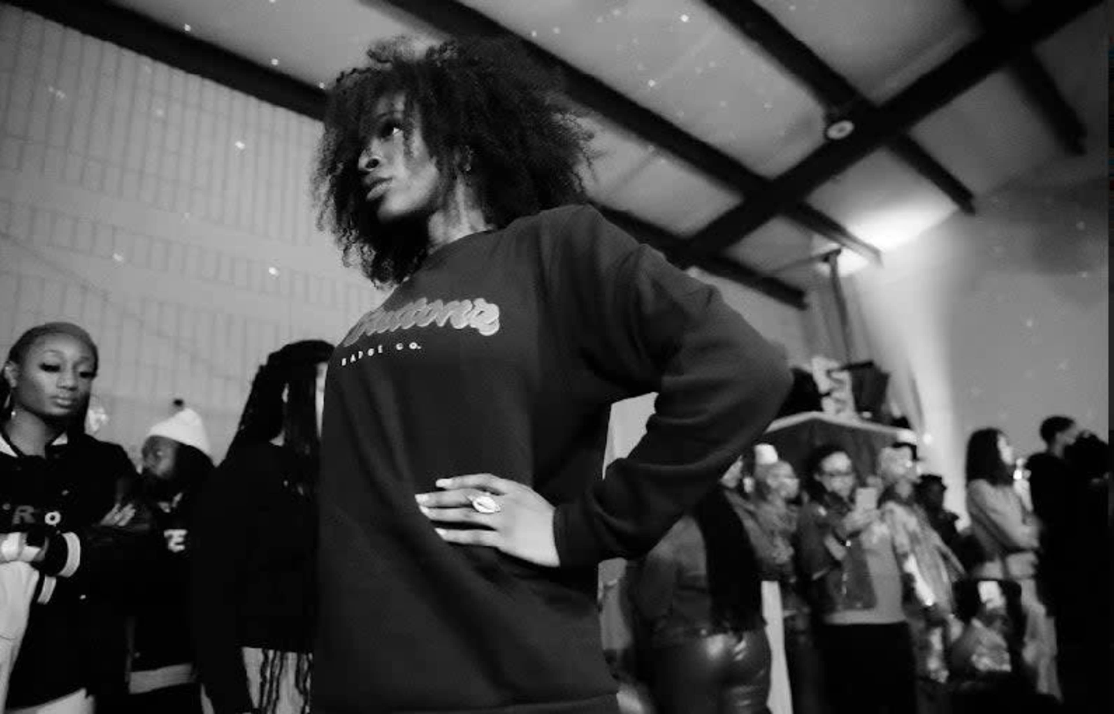 A model poses confidently in a black sweatshirt while spectators gather in the background.