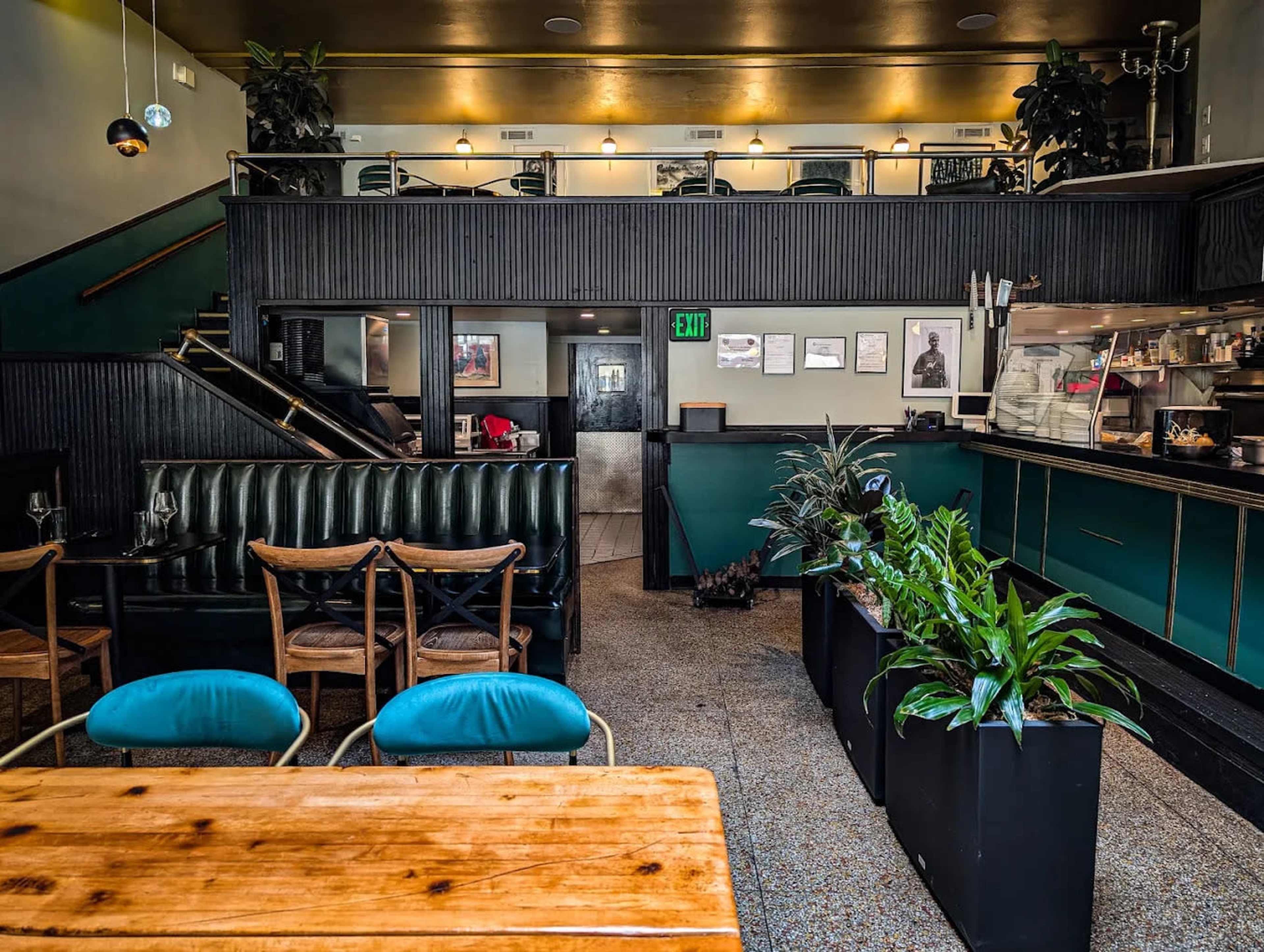 The image shows a stylish restaurant interior featuring a combination of dark wood accents, green walls, potted plants, and a bar area with framed photographs.