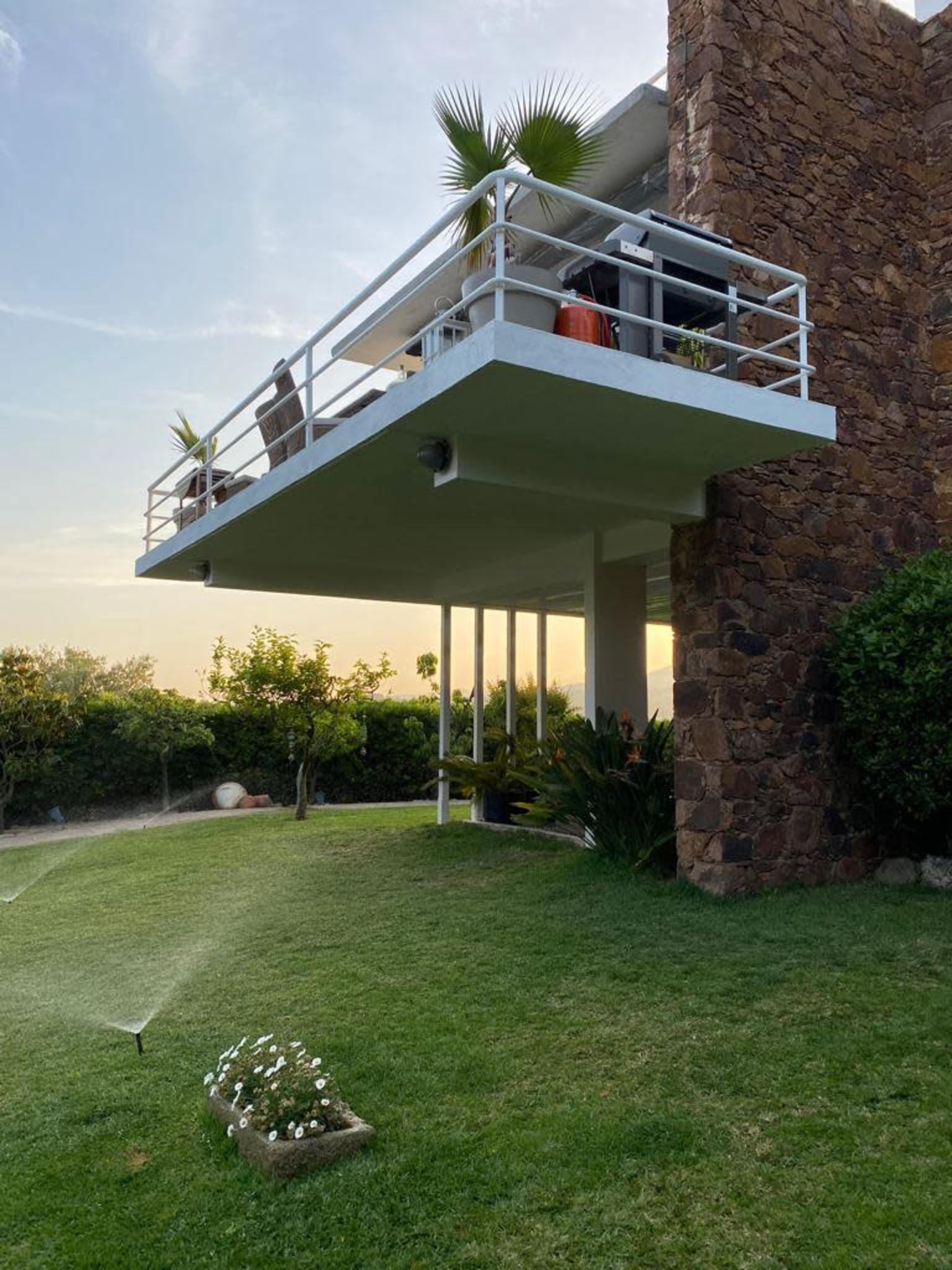 A modern house features a cantilevered balcony with potted plants overlooking a green lawn, where sprinklers water the grass.