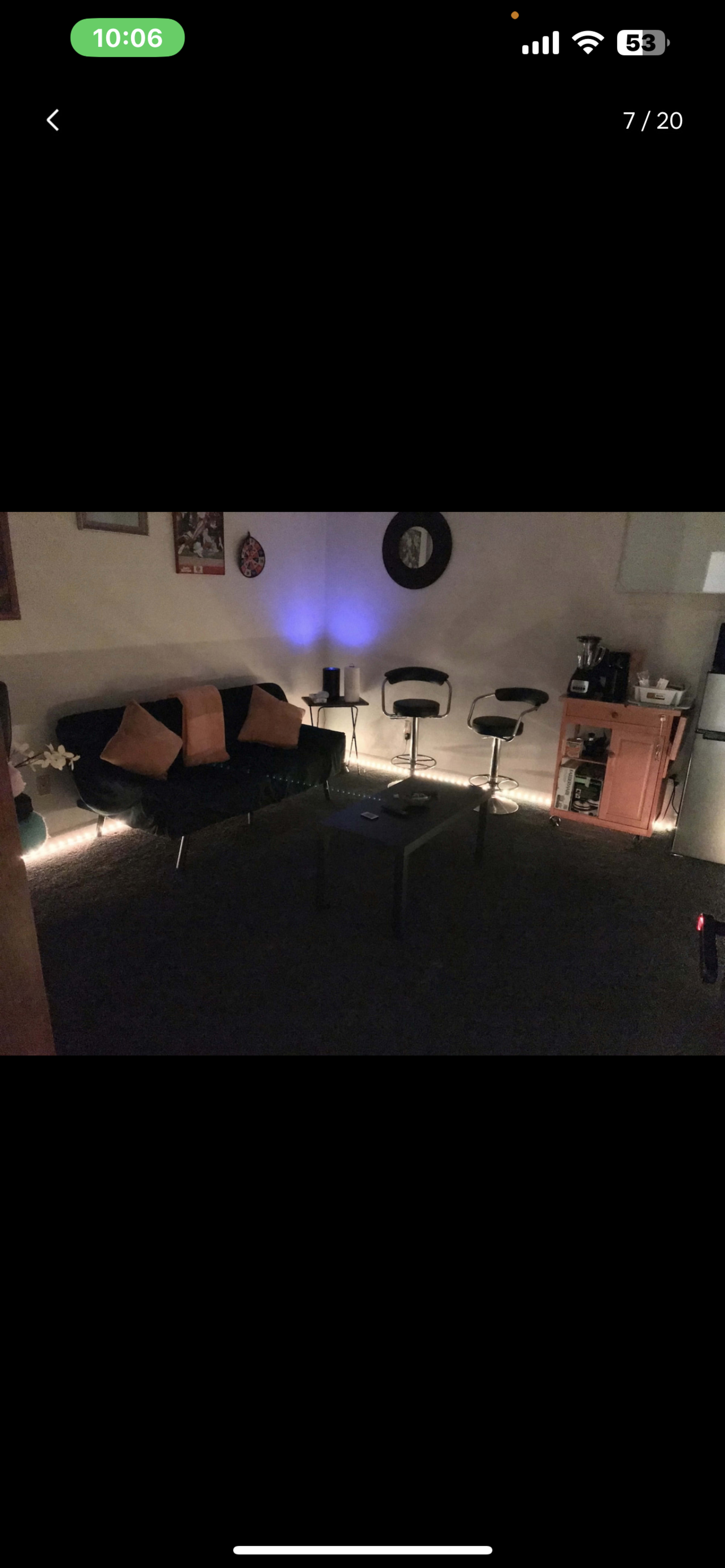 The image shows a dark living area with a black couch, four pillows, a coffee table, two bar stools, and a kitchenette visible in the background.