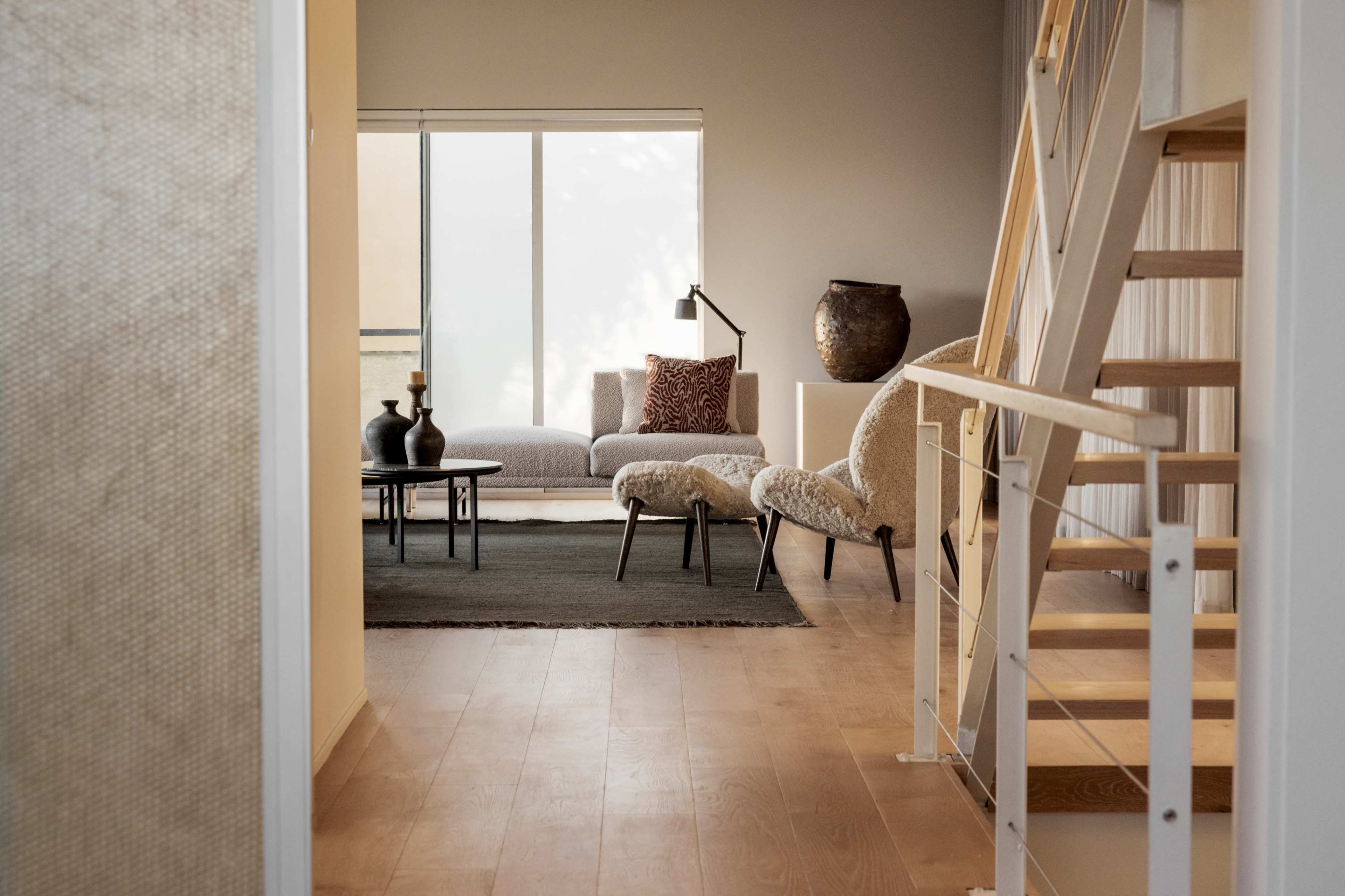 A staircase leads to a modern living room furnished with a sofa, armchair, and decorative items, illuminated by natural light from a large window.