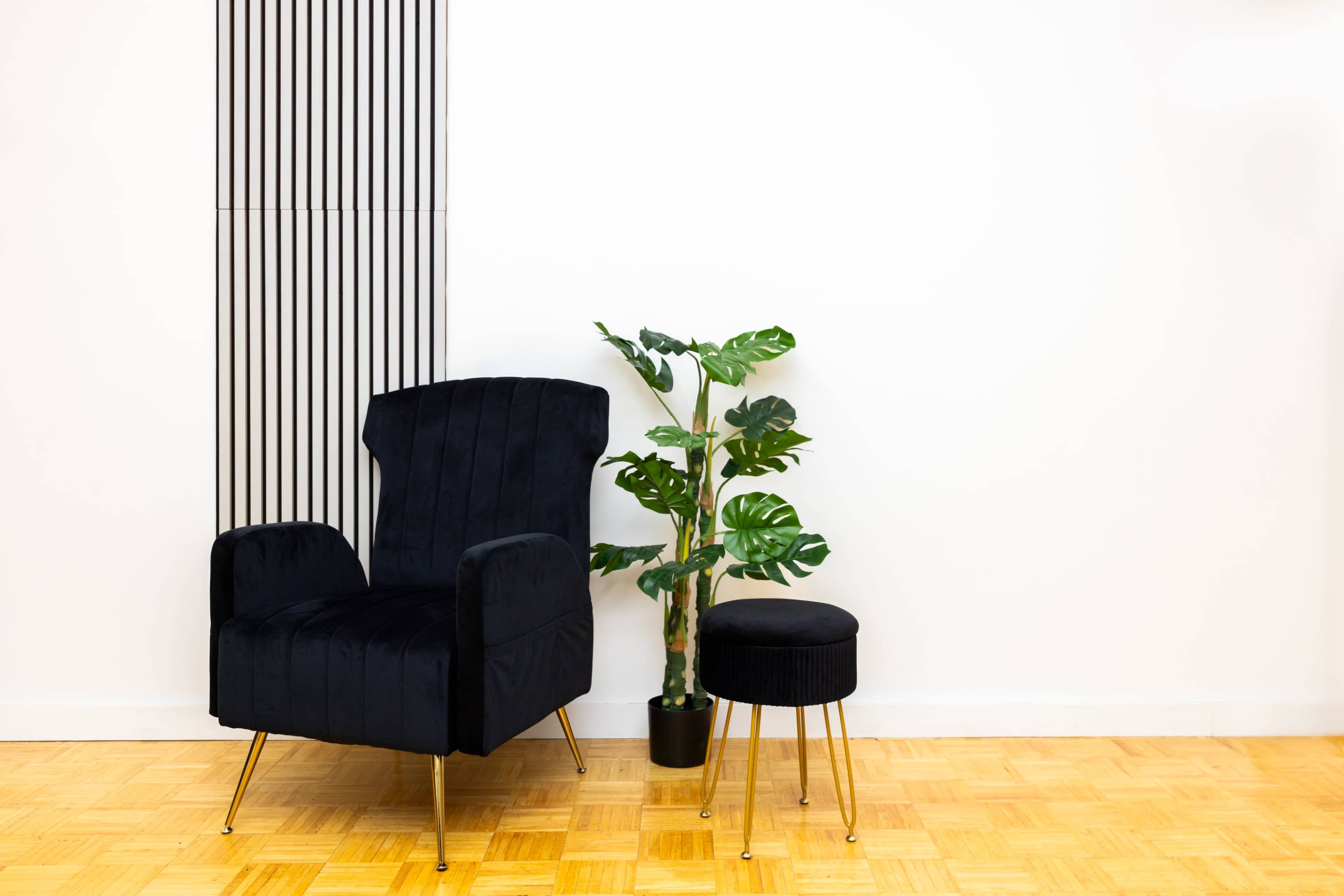 A black upholstered chair and a small matching stool are positioned next to a potted plant against a white wall with vertical stripes.