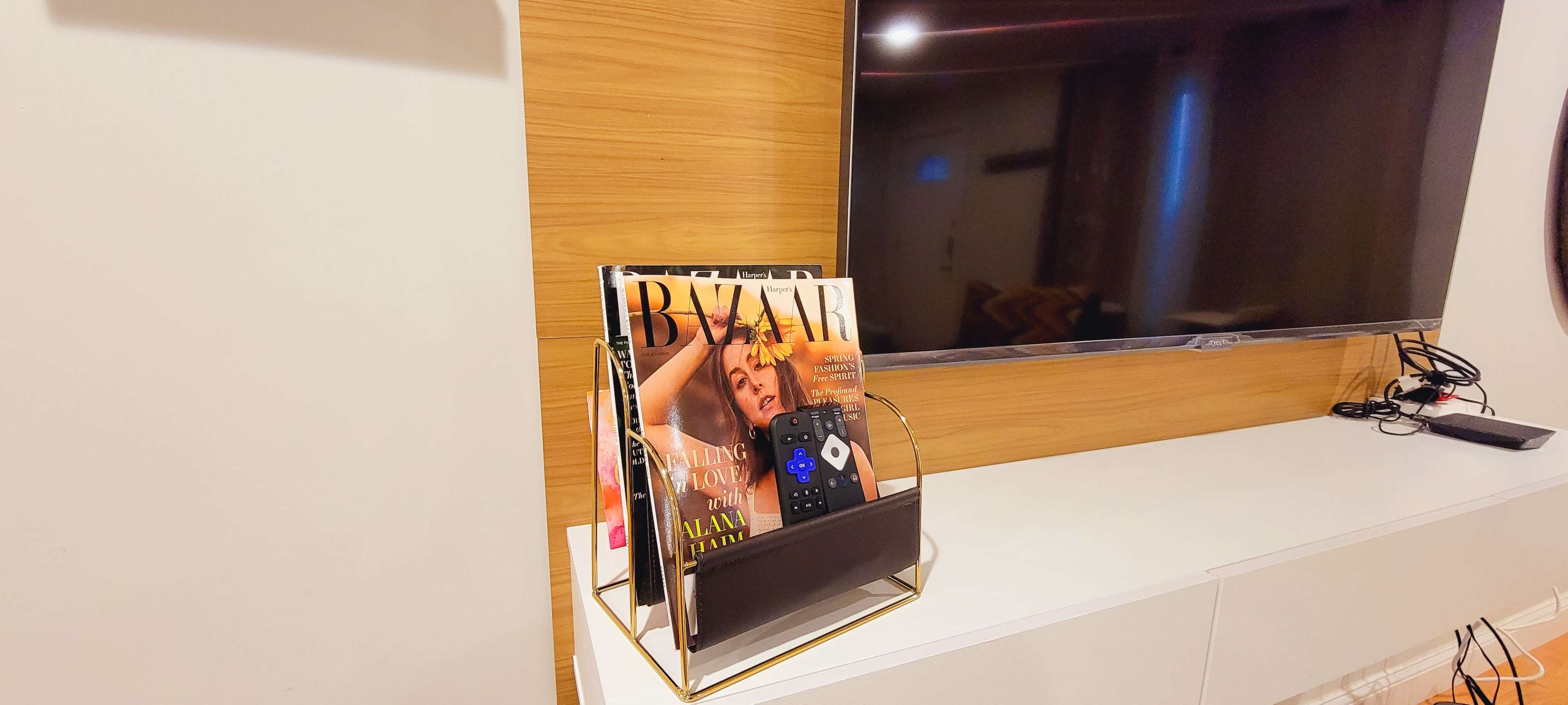 A magazine rack holds several issues of Harper's Bazaar and a remote control beside a television on a white cabinet.