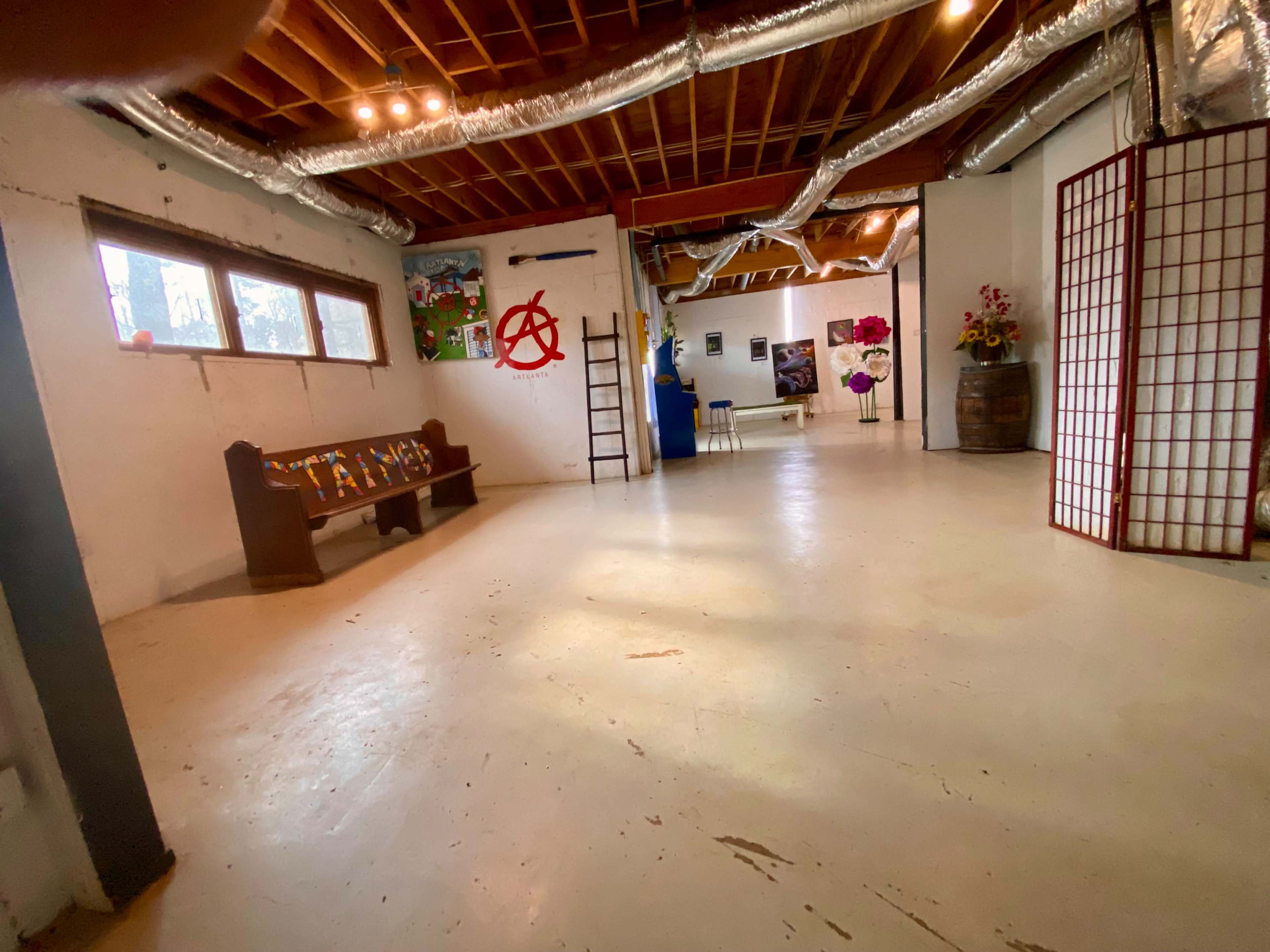 The image shows an open space with a concrete floor, featuring a bench, artwork on the walls, and a few decorative items.
