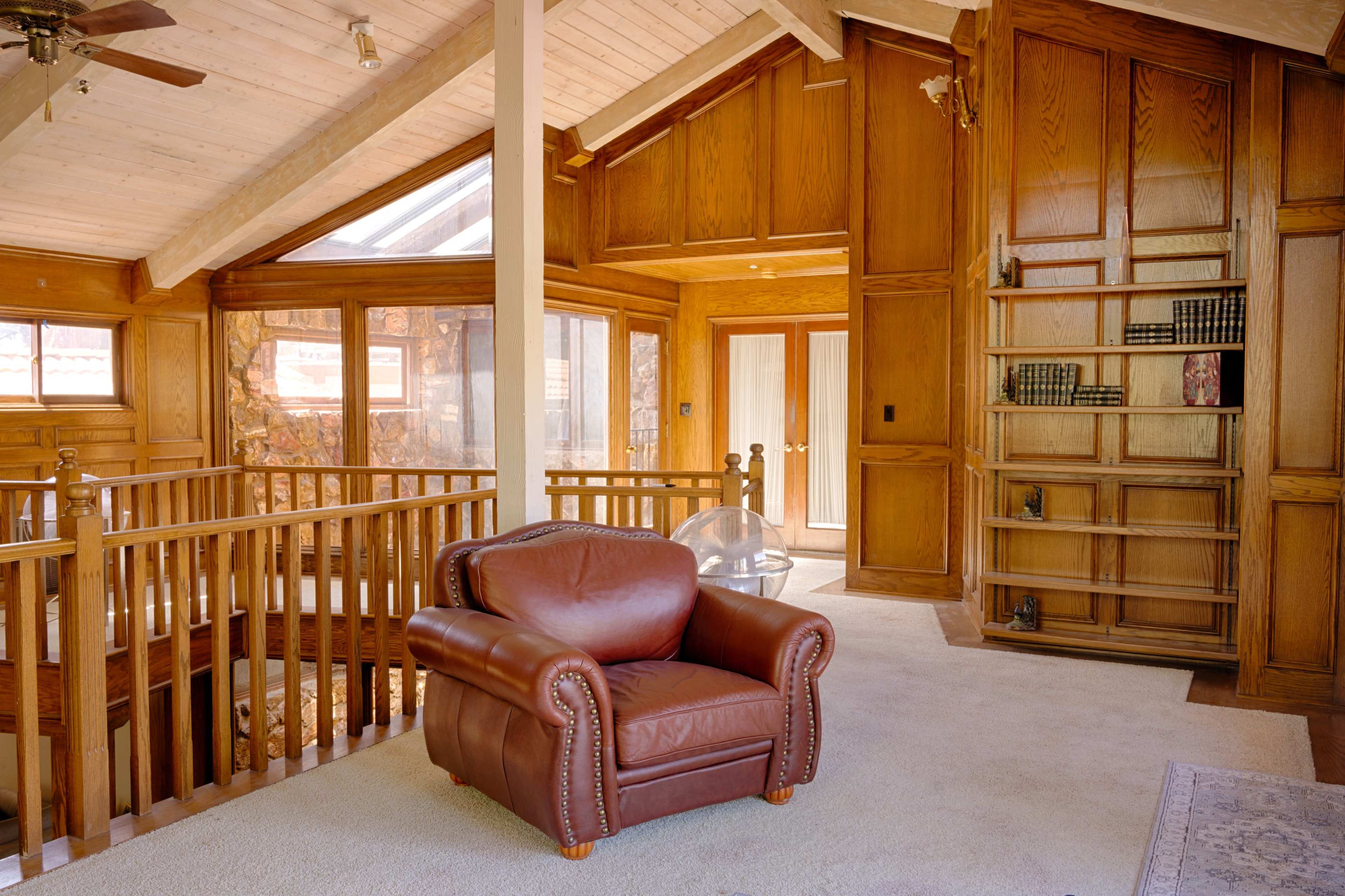 The image shows a spacious, wooden-paneled interior with a plush brown armchair, a balcony railing, and a built-in bookshelf.