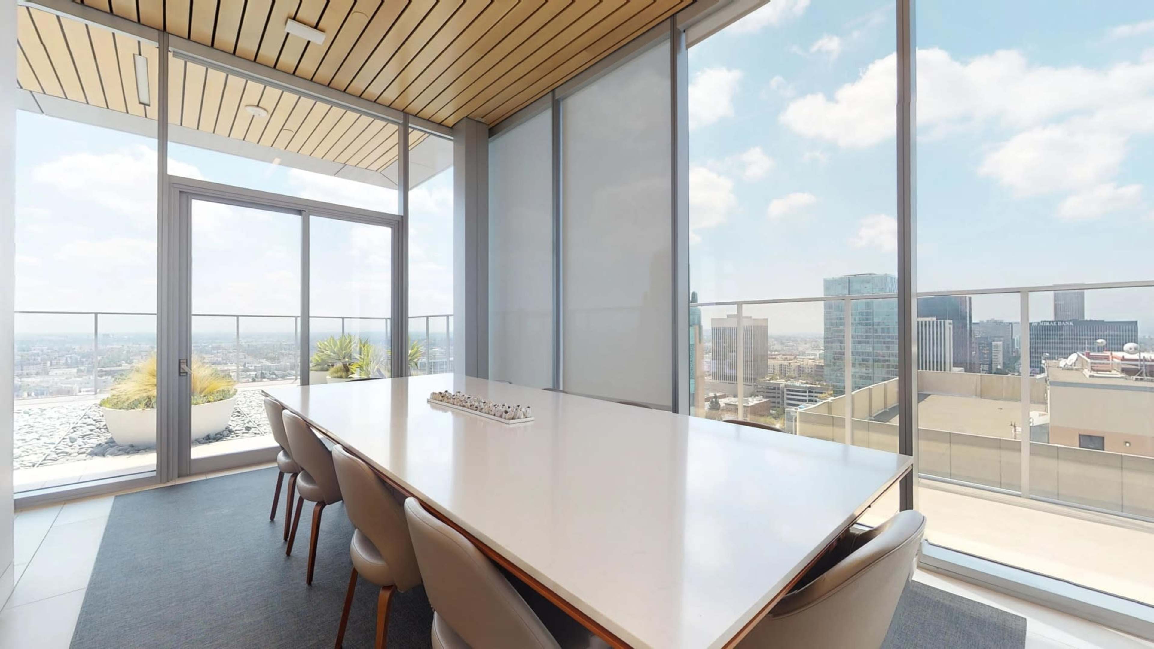 The image shows a modern conference room with a large white table and panoramic windows overlooking a city skyline.