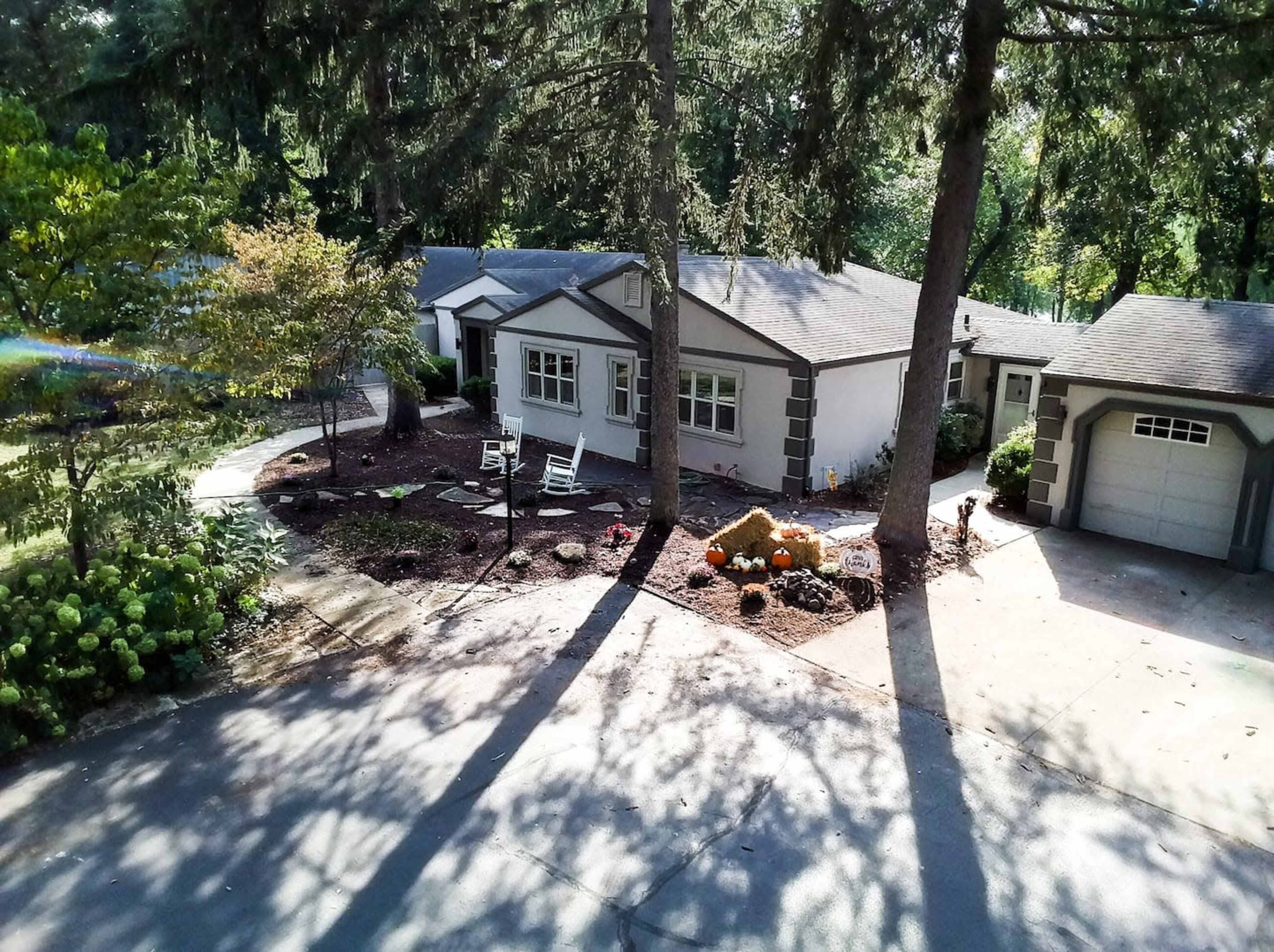 A view of a gray house surrounded by landscaped gardens, featuring a driveway and several trees.