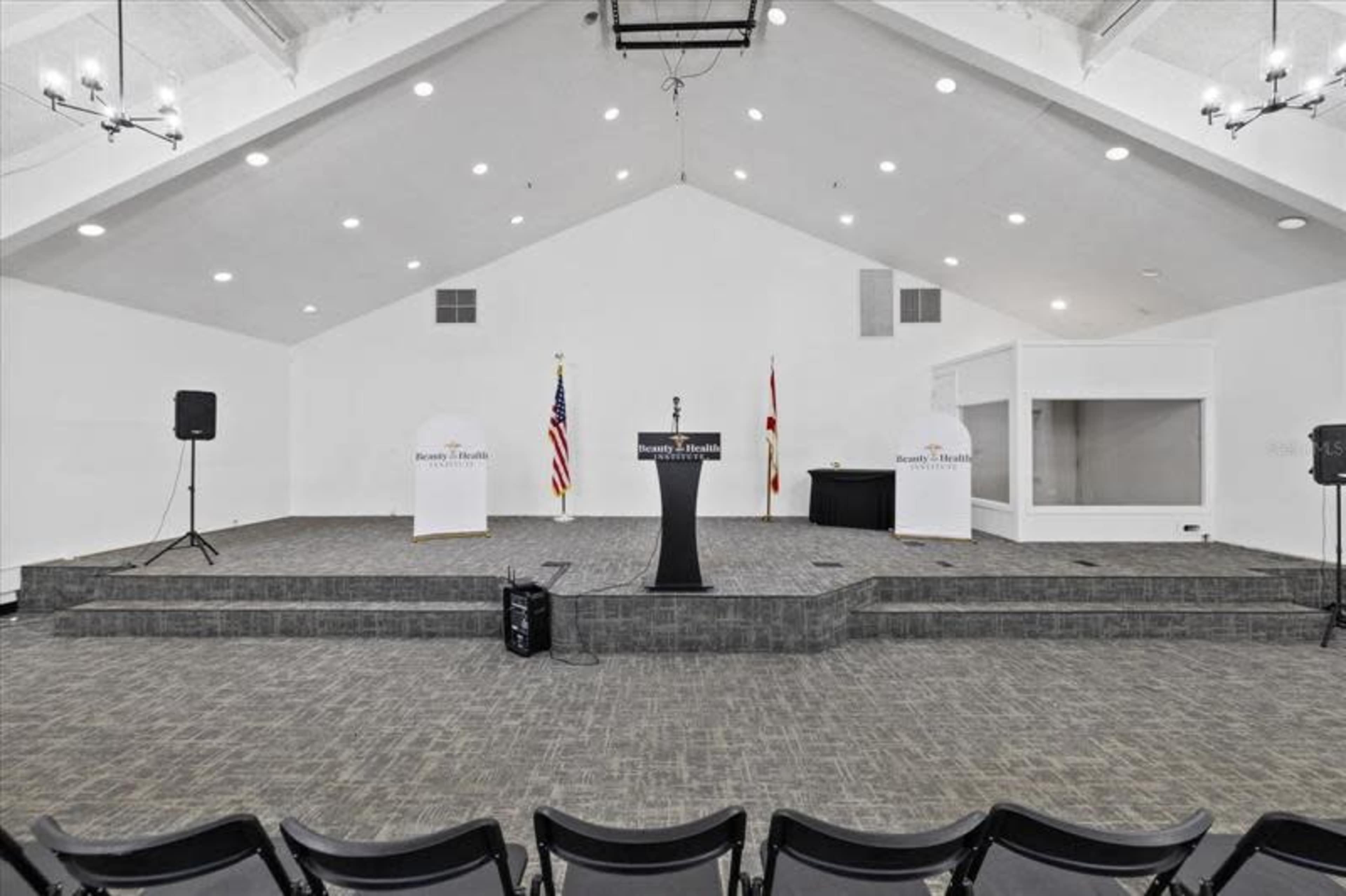 The image shows an empty stage setup for an event, featuring a podium with a microphone, U.S. and state flags, and seating arranged in front of it.