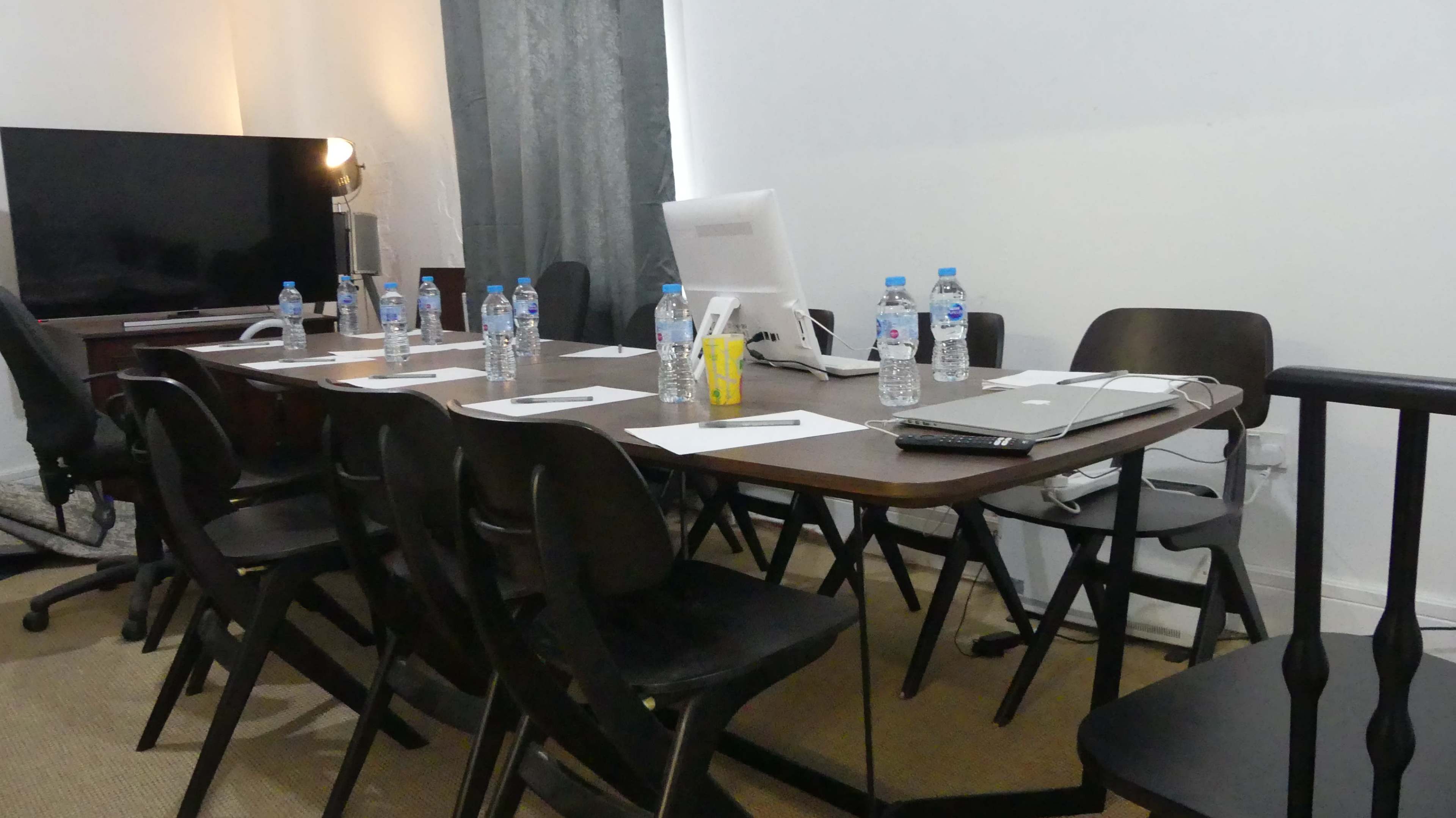 A rectangular table is set up in a room with several chairs around it, featuring water bottles, papers, and a computer monitor.