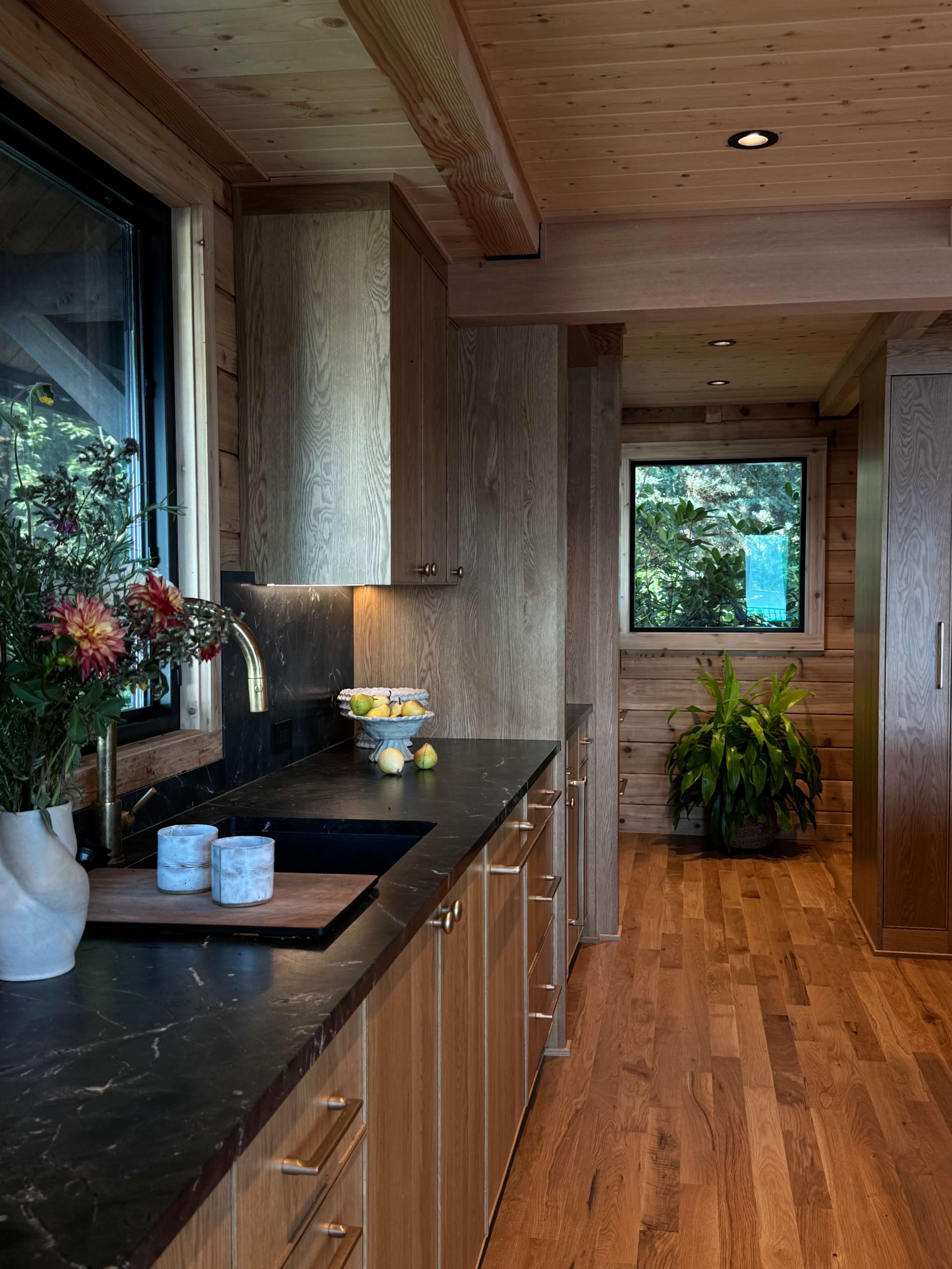 A modern kitchen features wooden cabinetry, a dark countertop, and a window with greenery visible outside.