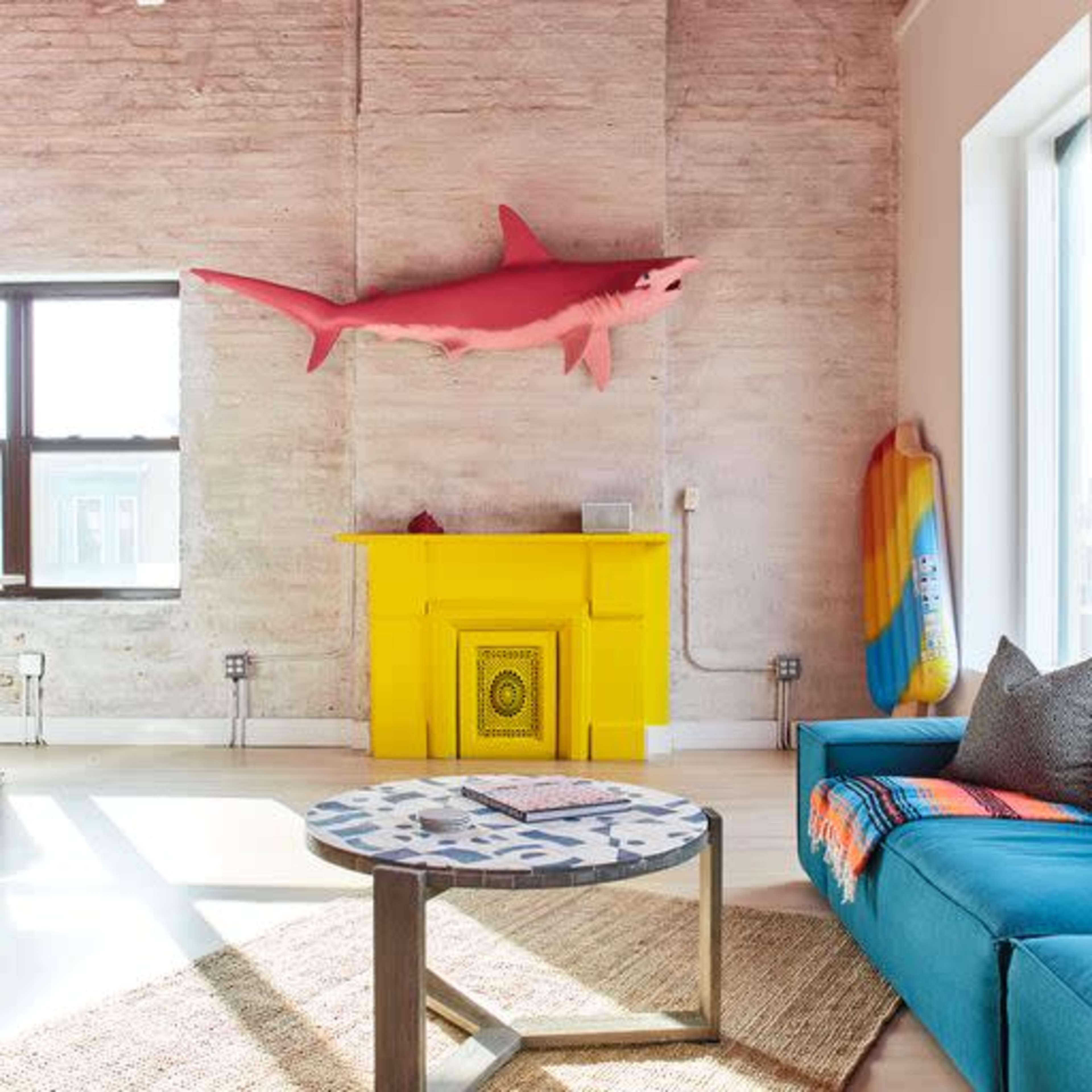 A bright, modern living room with a blue sofa, a patterned coffee table, a yellow cabinet, and a shark decoration mounted on the wall.