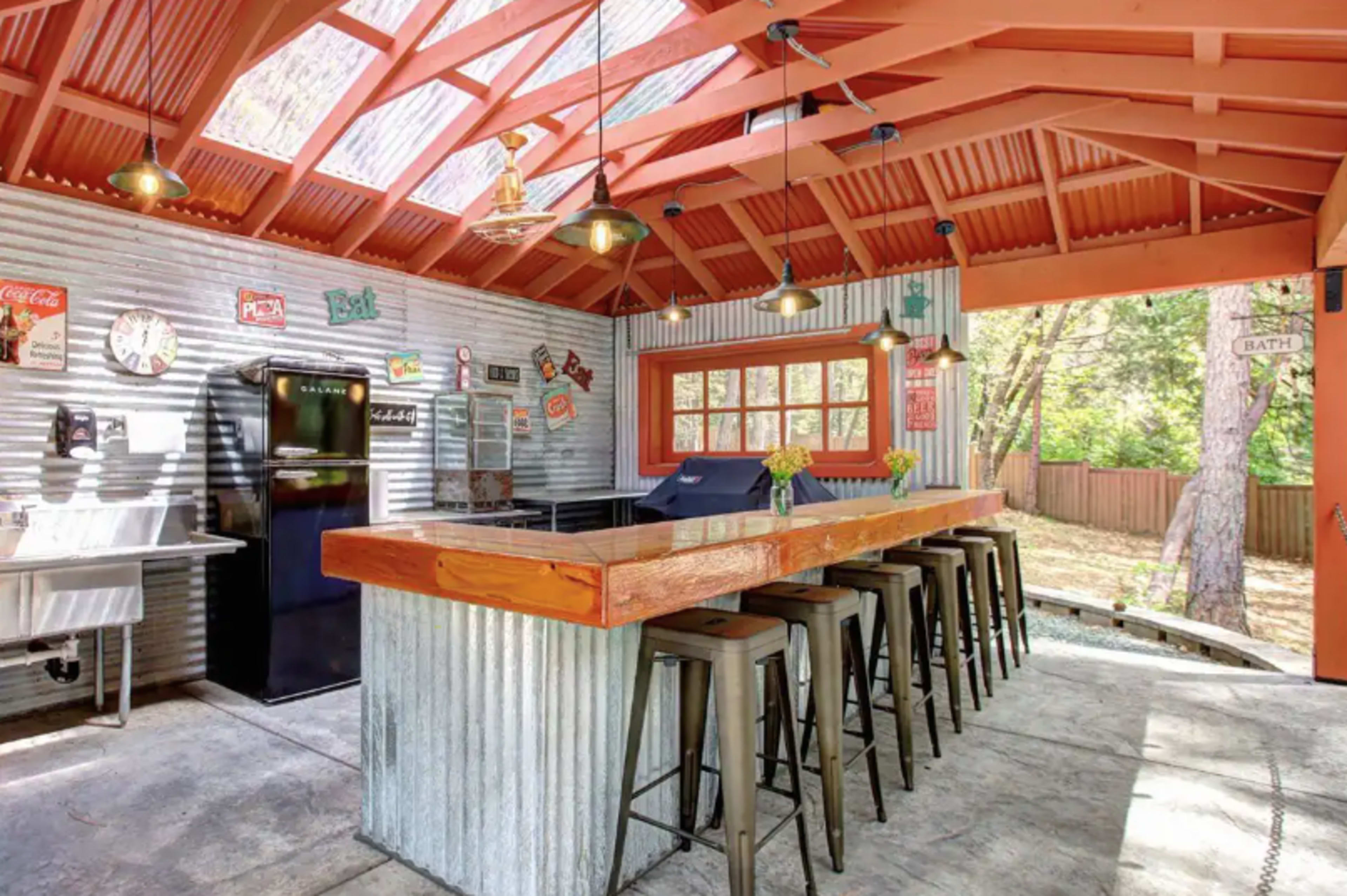 A rustic outdoor kitchen with a wooden bar, metal stools, and a corrugated steel backdrop, all under a slanted roof with ample natural light.