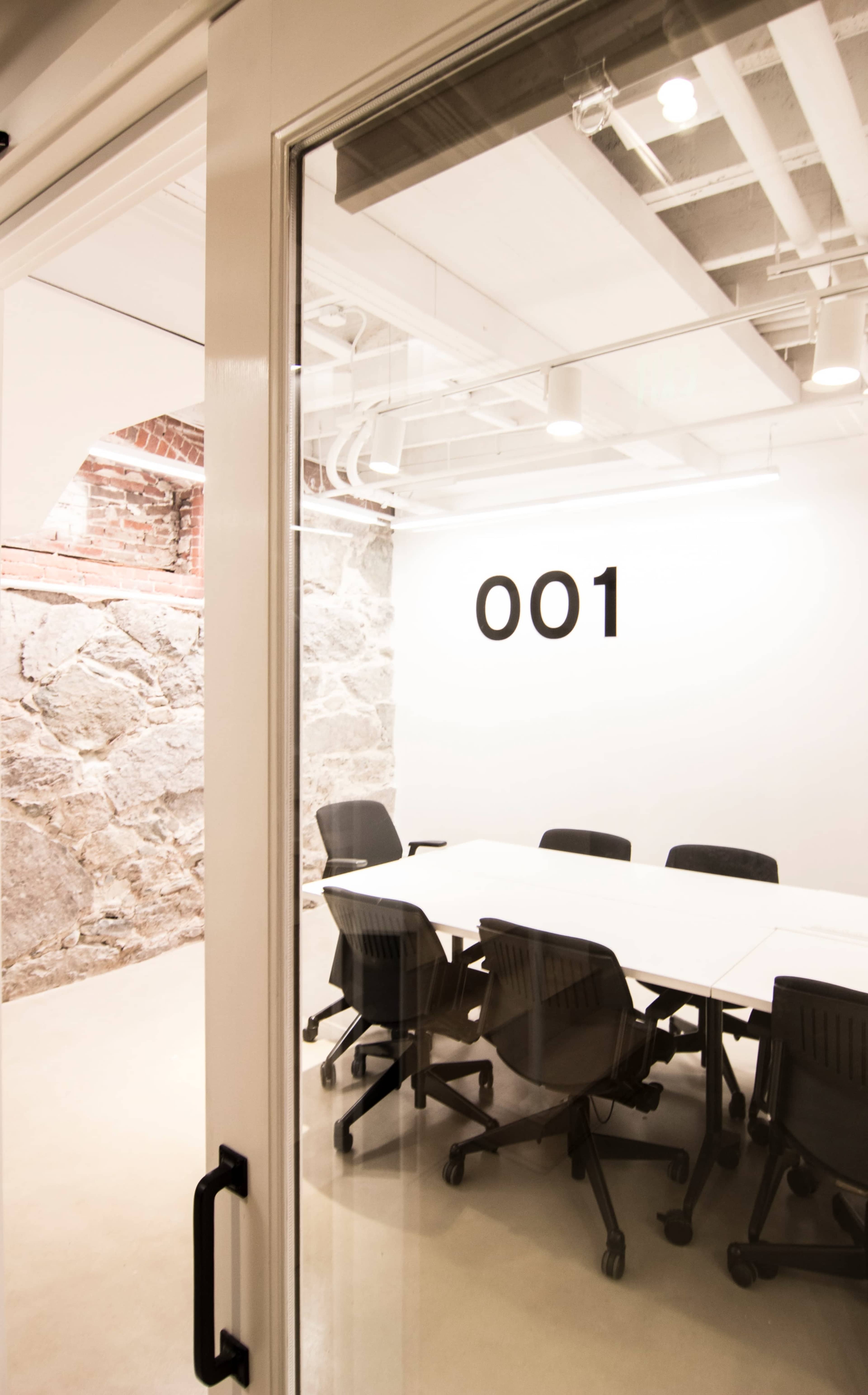 A conference room with a large white table and black chairs is visible through a glass door, featuring a wall with the number "001" and exposed brick.