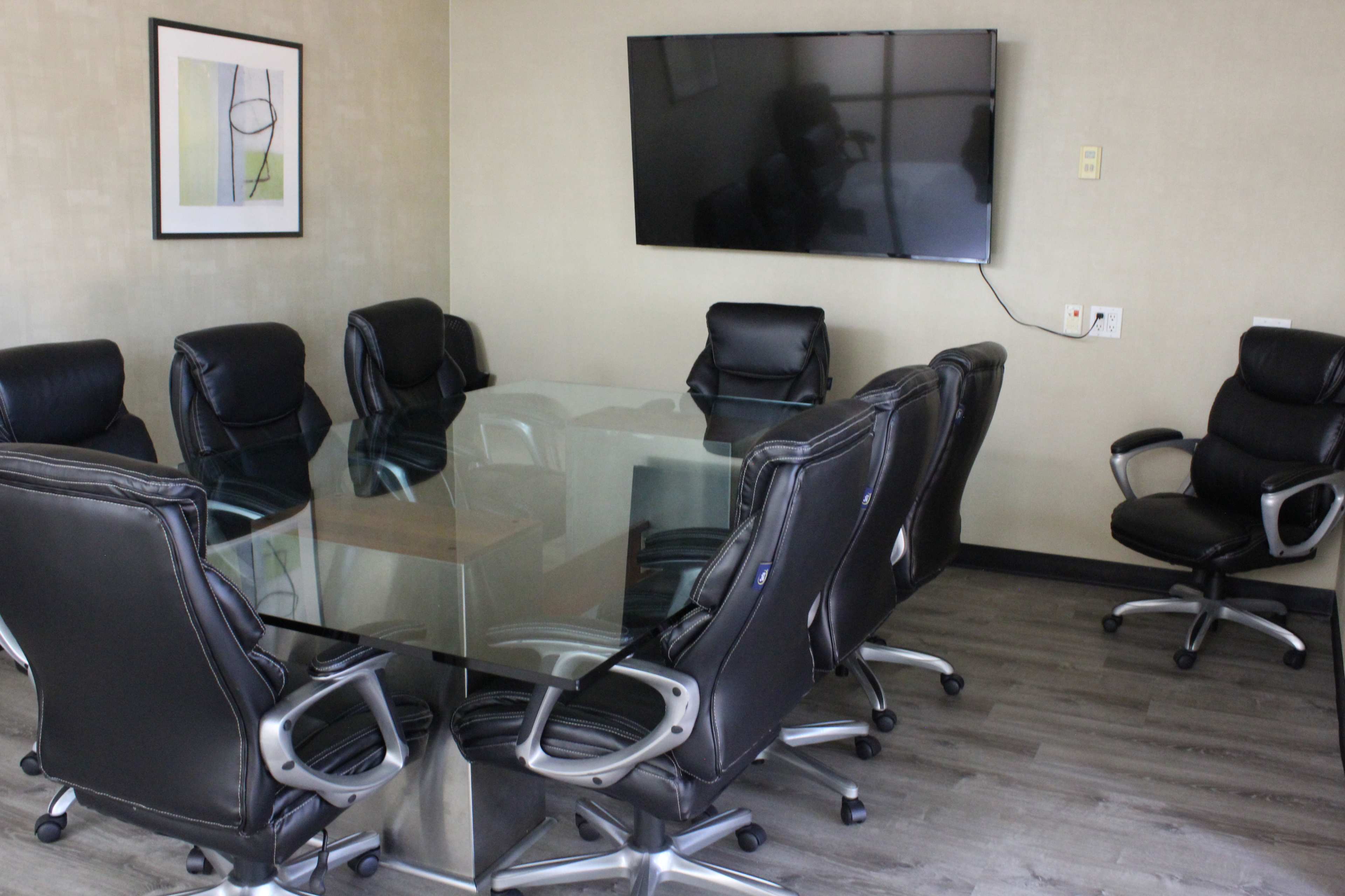 A modern conference room features a glass table surrounded by eight black leather chairs and a mounted television on the wall.