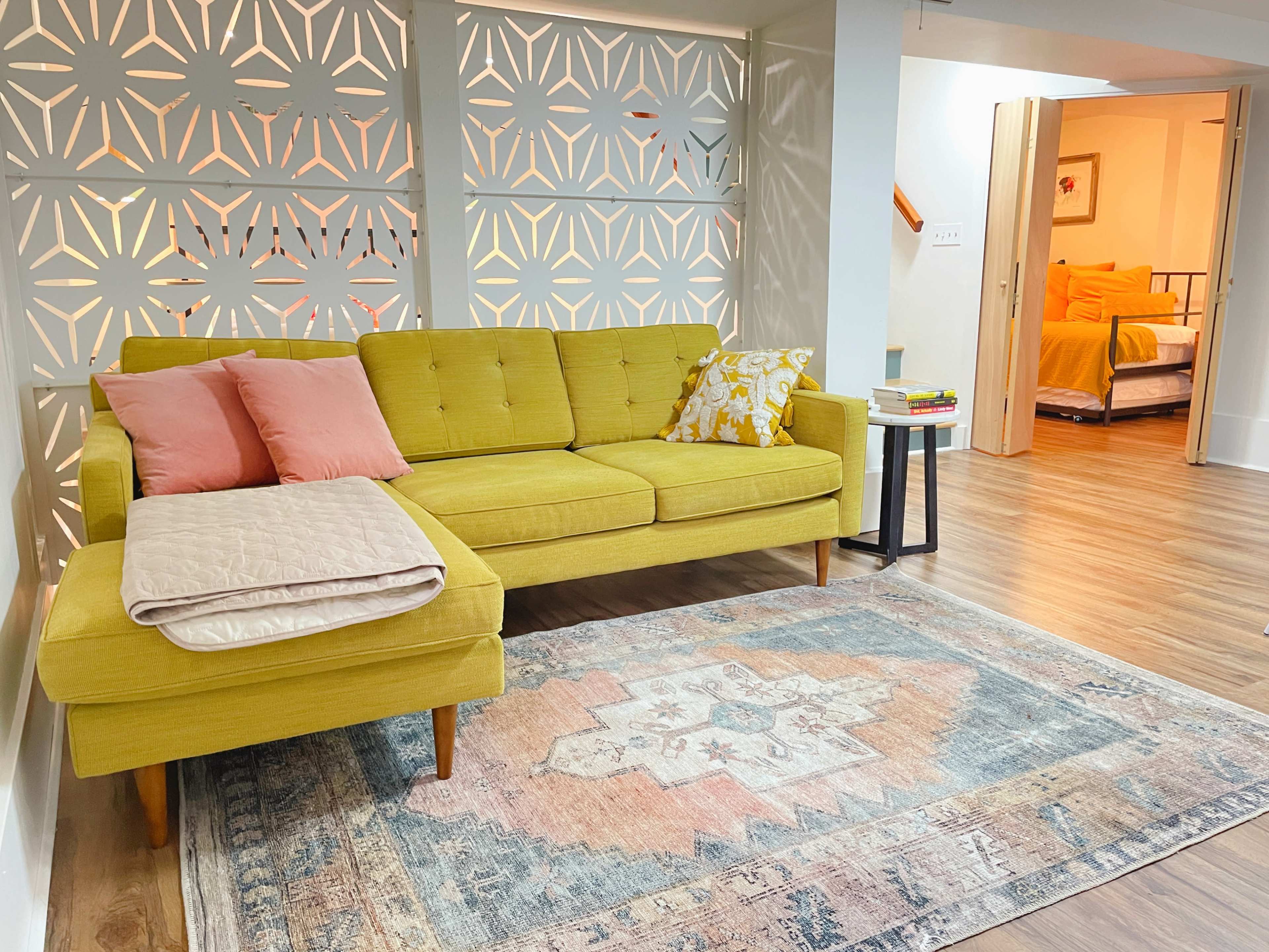 A mid-century modern yellow sectional sofa with pink and patterned cushions, placed on a patterned area rug in a spacious room with decorative partition screens and a glimpse of a bedroom in the background.