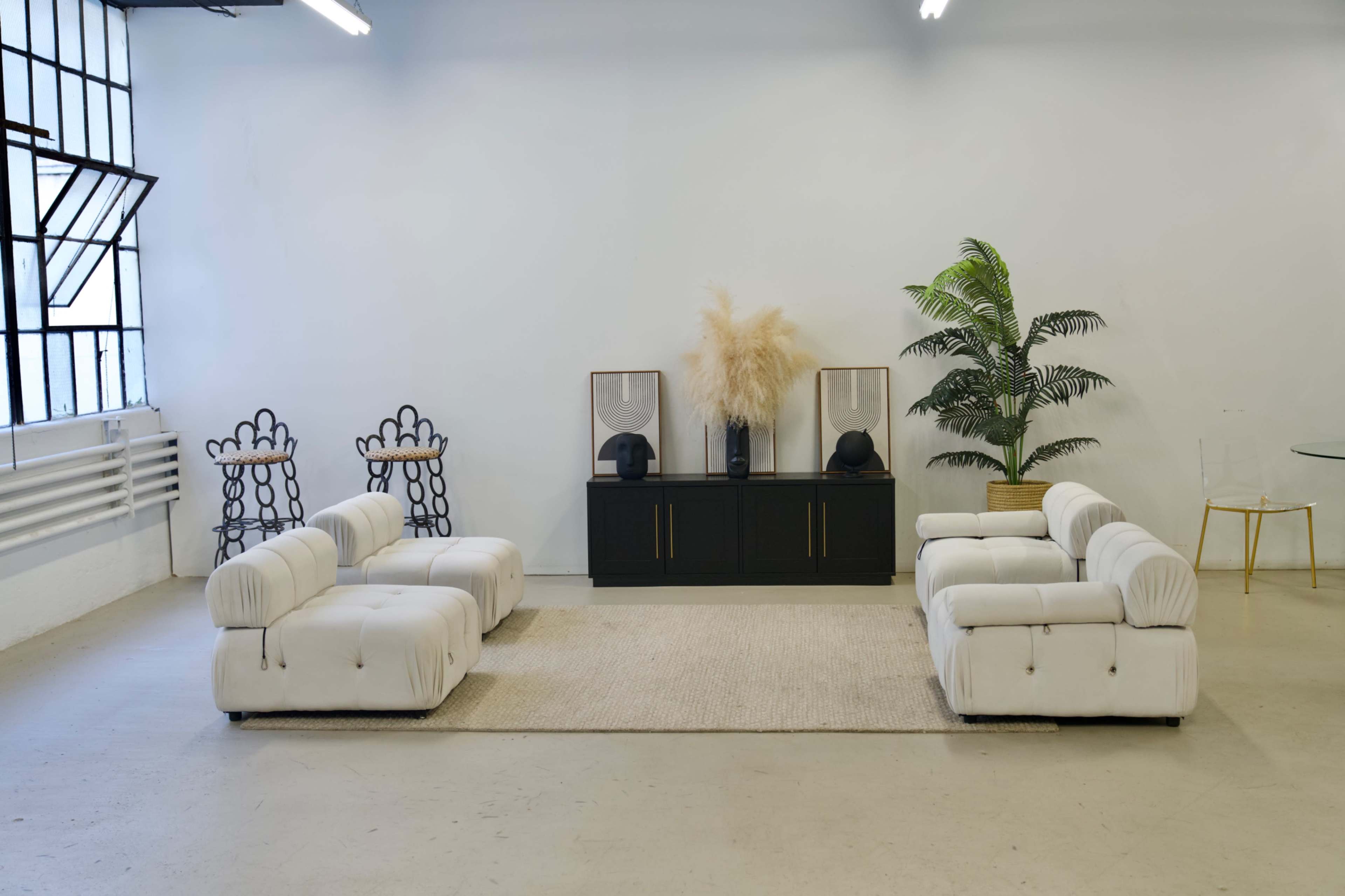 A minimalist interior space featuring two white sofas facing each other, a black cabinet with decorative items, and a large potted plant against a white wall.