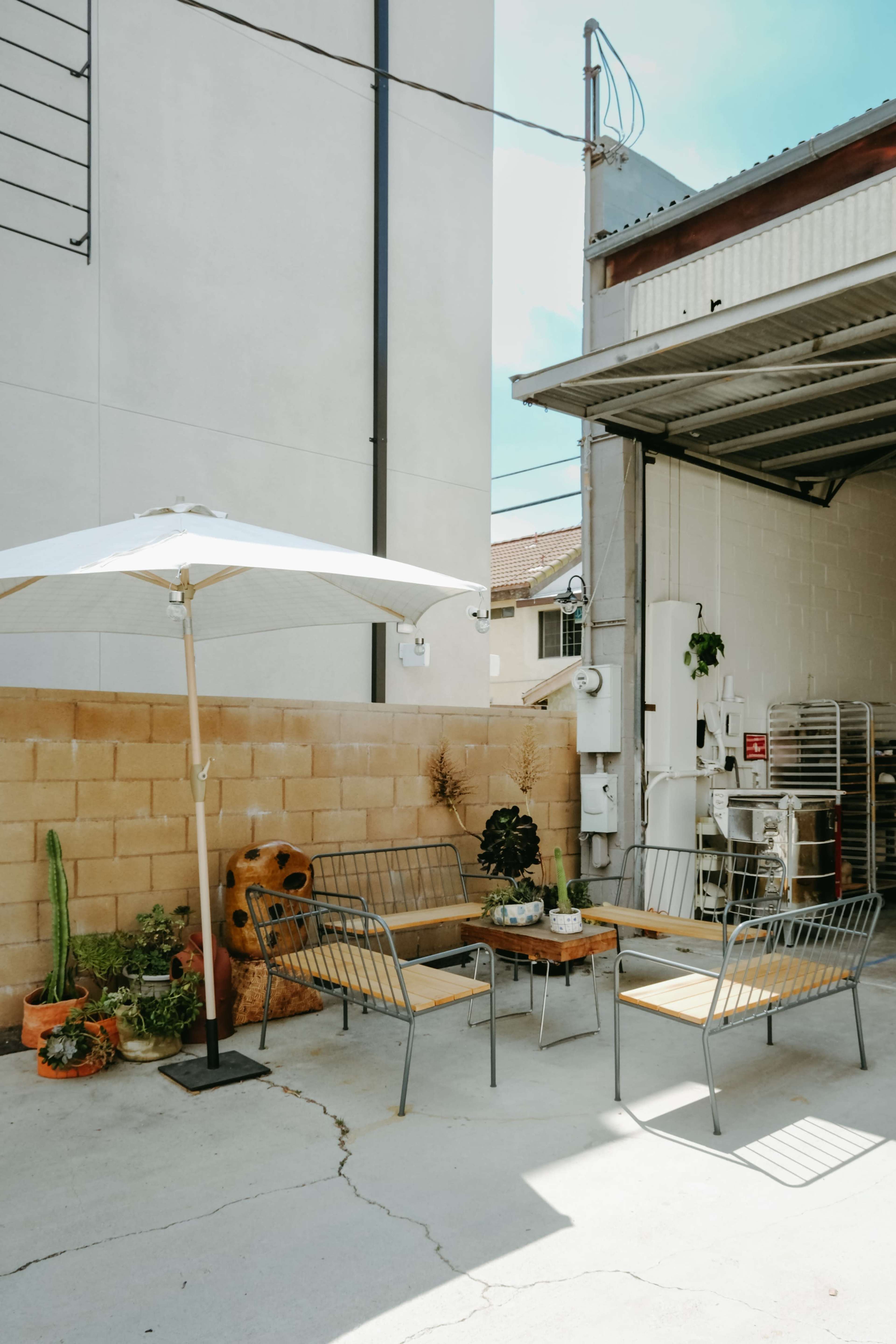A small outdoor seating area featuring metal chairs and a table, shaded by a large umbrella, against a concrete wall with various plants.
