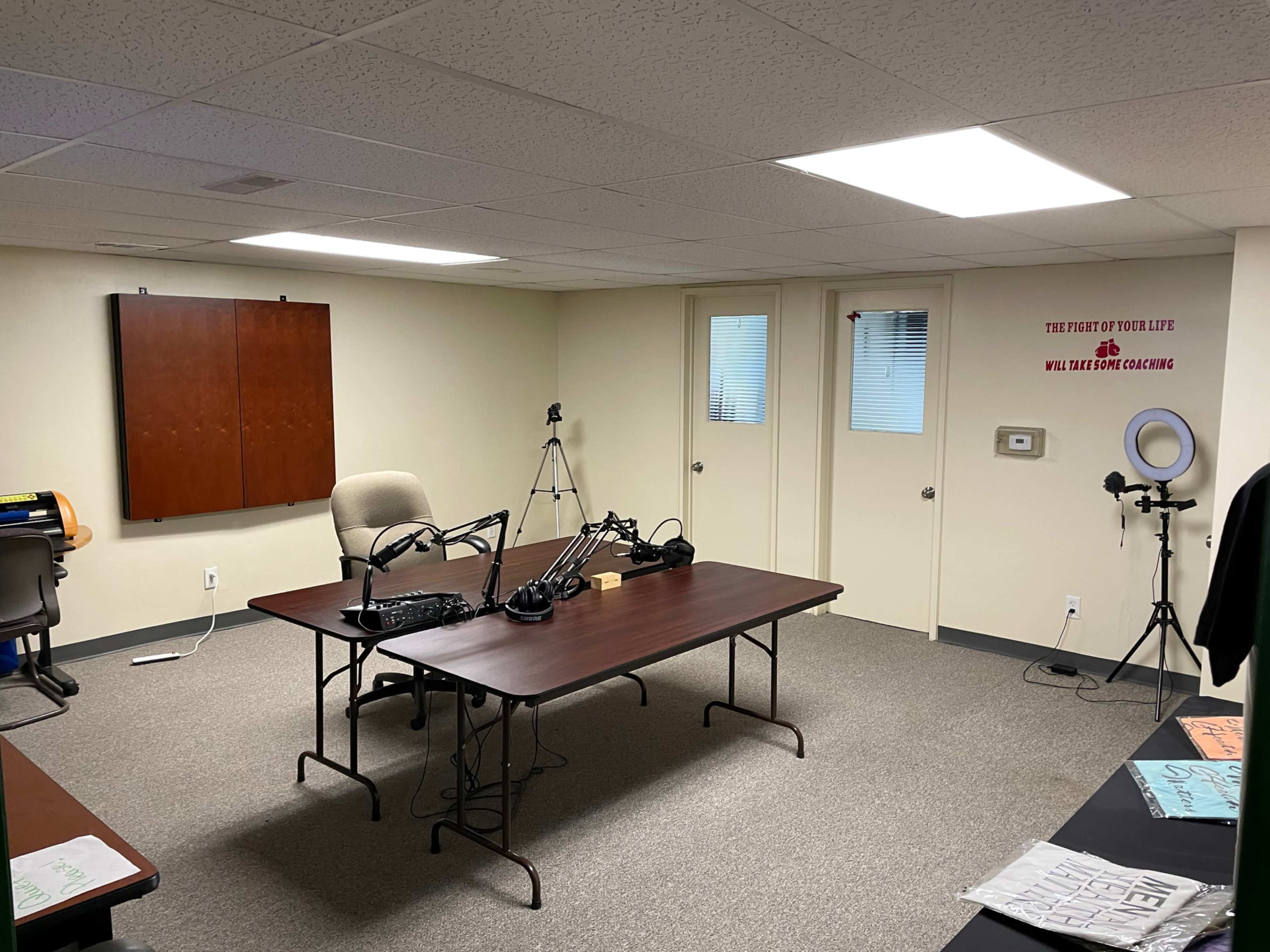 The image shows a recording studio with a large table, microphone setups, and a tripod in a sparsely furnished room.