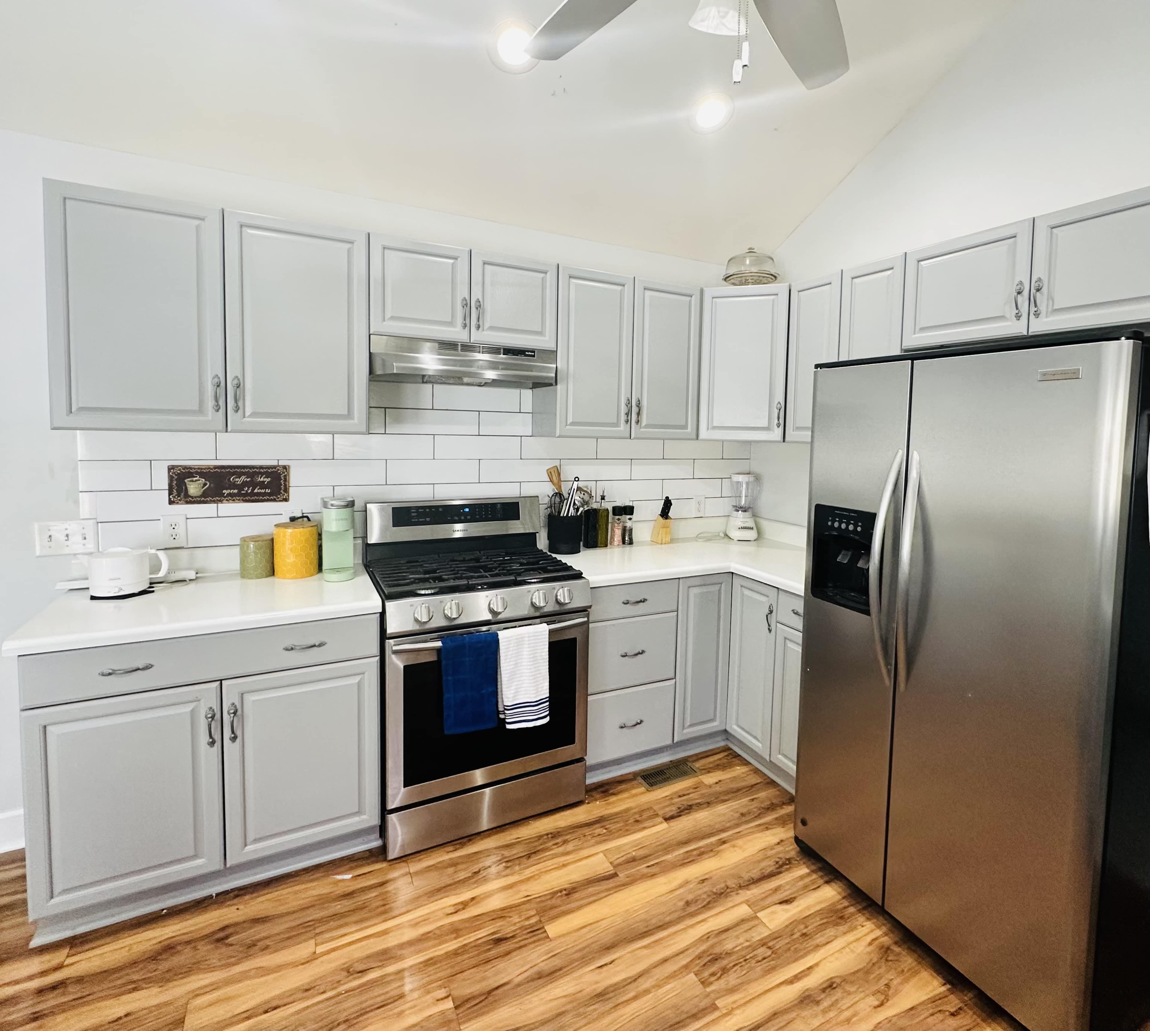The kitchen features gray cabinets, stainless steel appliances, and a white tile backsplash, with wooden flooring.